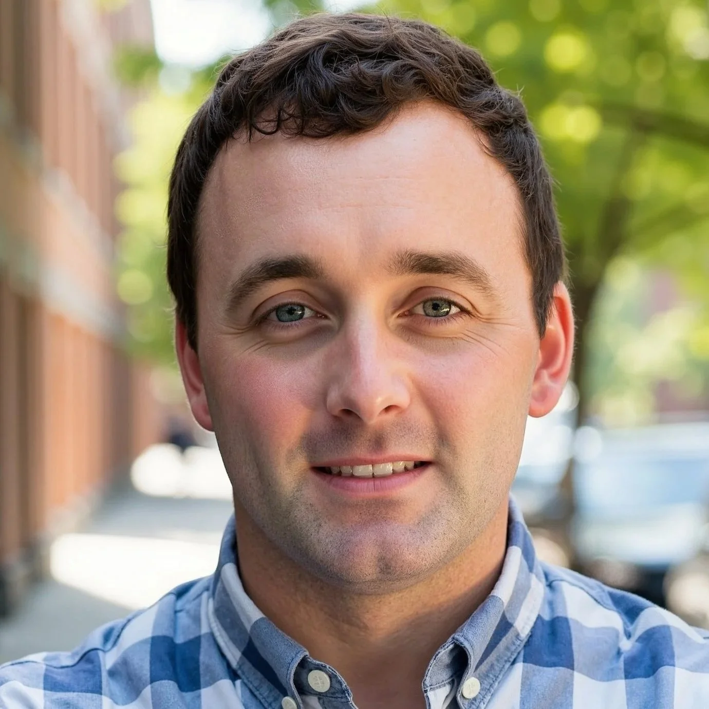 Close-up photo of a smiling man with short dark hair, light blue eyes, and wearing a blue and white checkered shirt outdoors during daytime.