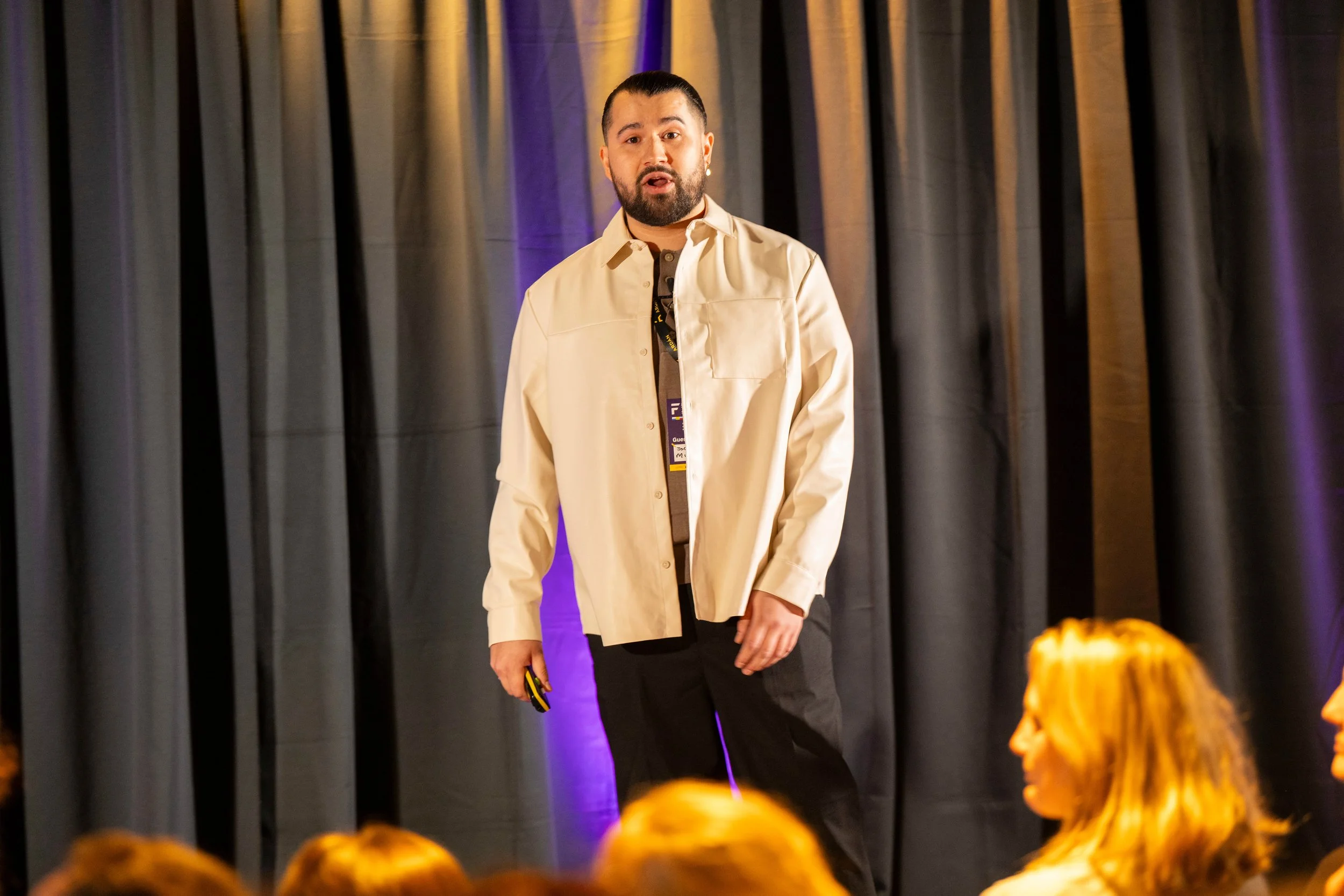 A man with a beard and bun hairstyle, wearing a cream-colored shirt over a dark shirt, is speaking on stage in front of a dark curtain backdrop with purple lighting. There are audience members with orange hair visible in the foreground.