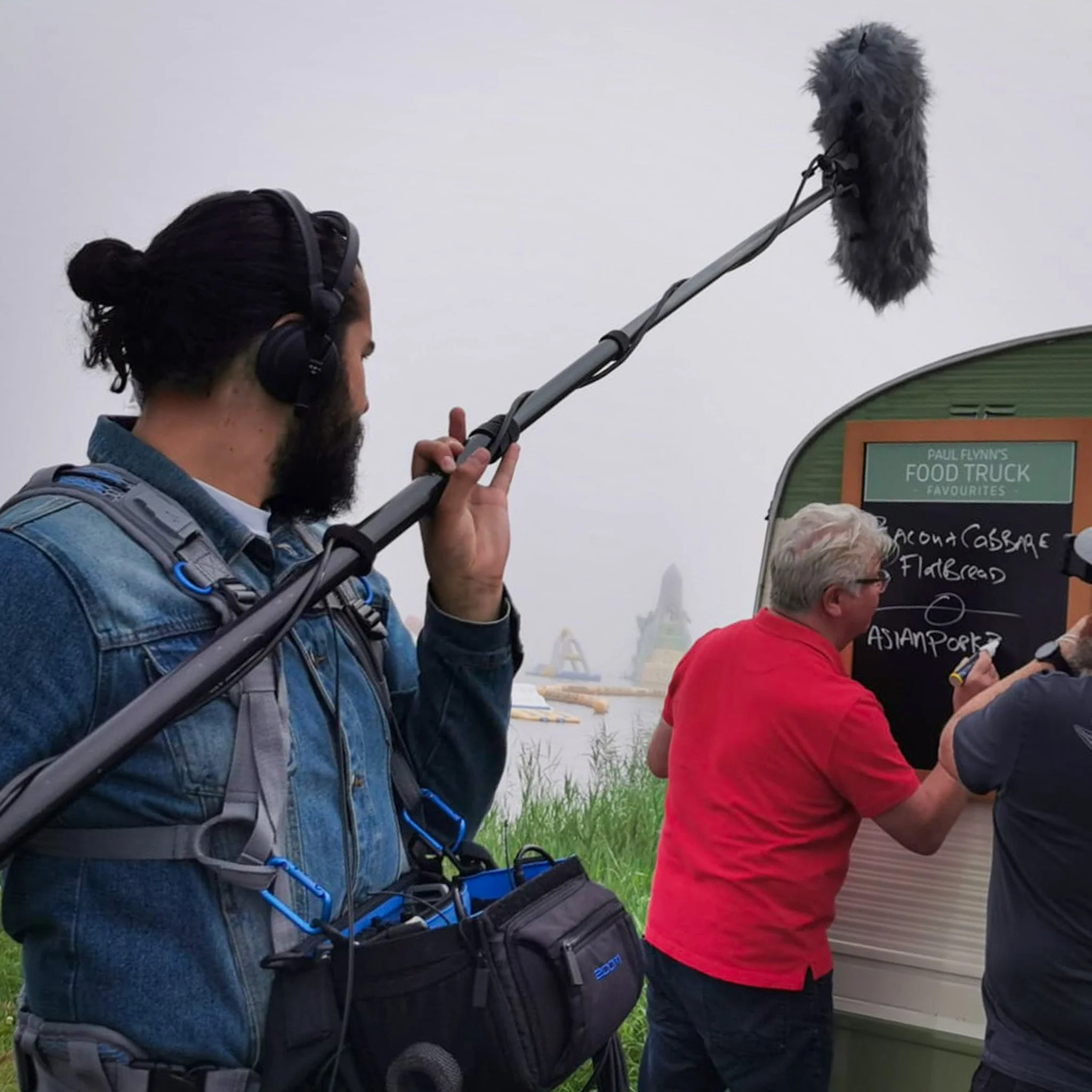 A man with dark hair and a beard holding a boom microphone with a furry windscreen at an outdoor event. The man is wearing headphones and a denim jacket. Other people are present, and there is a sign for a food truck.