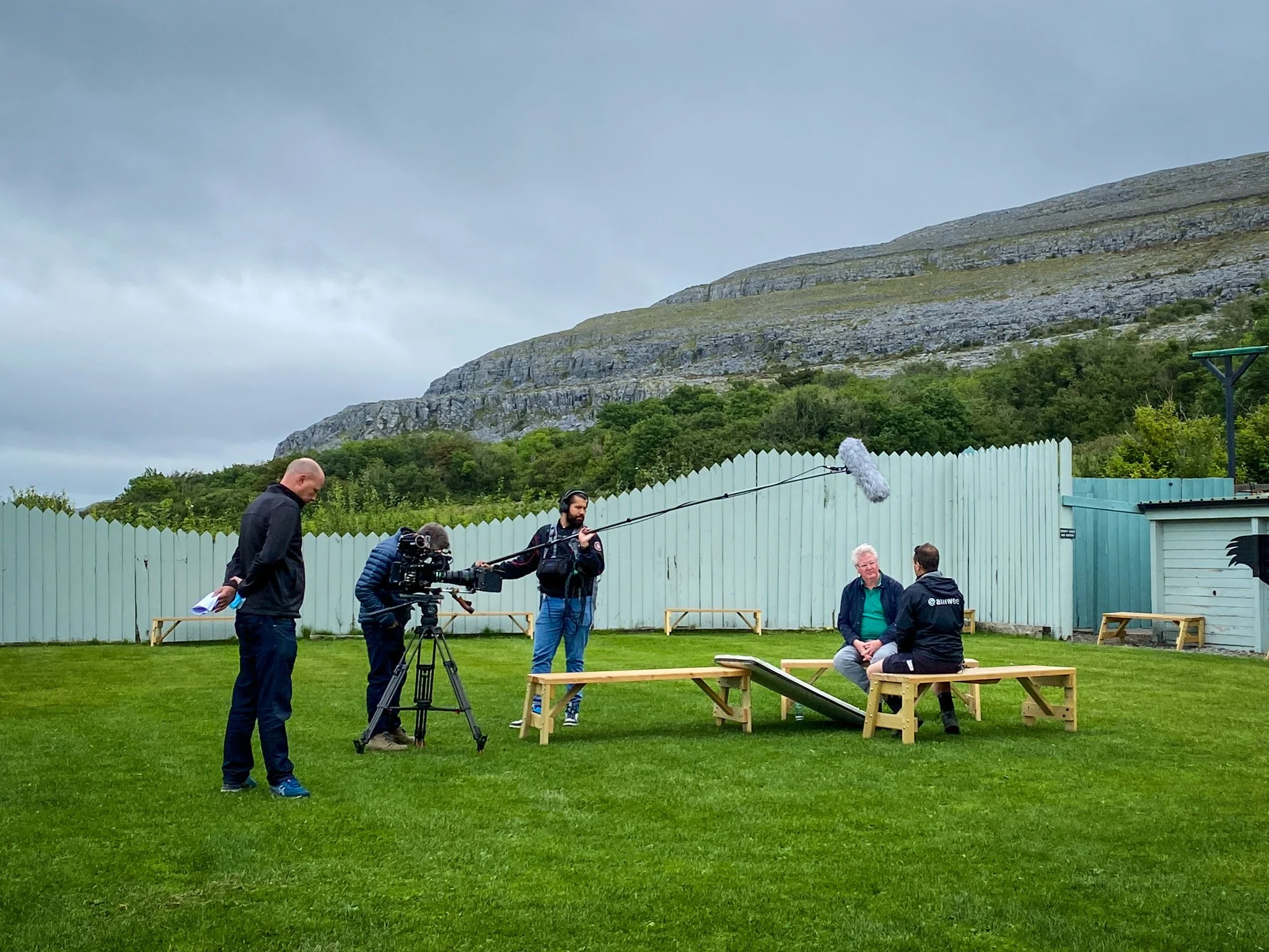 A film crew filming an interview or scene outdoors on a grassy area with wooden benches, green fence, and a mountain in the background. Crew members operate cameras and sound equipment, and some people are engaged in conversation.