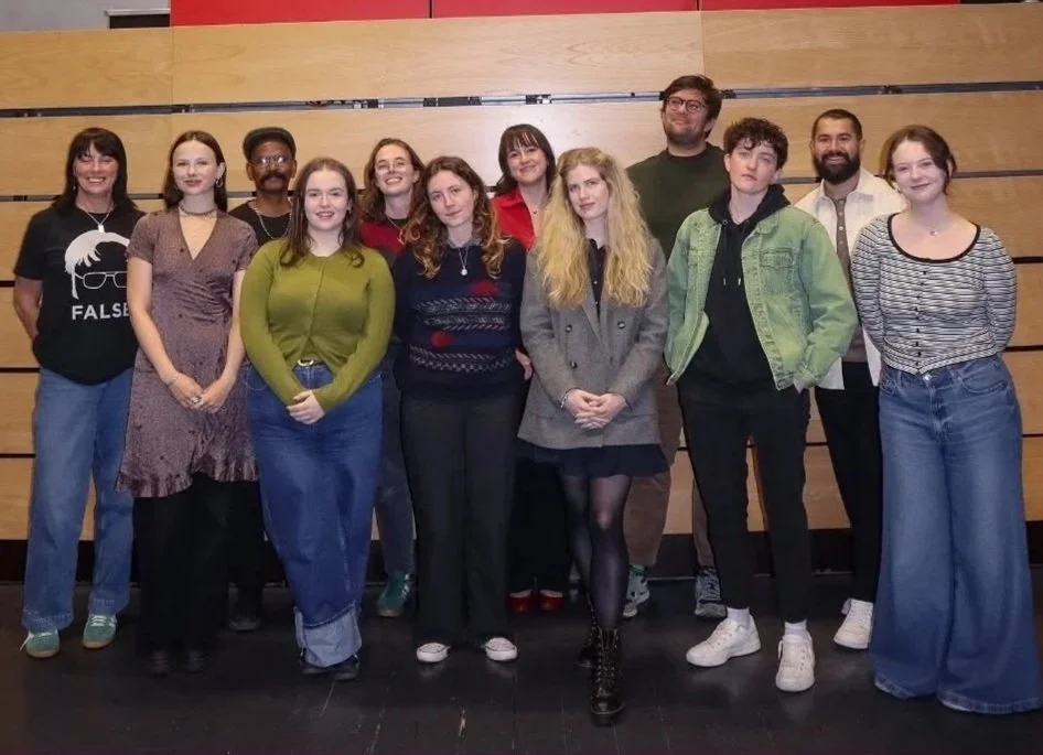 A group of twelve young adults standing together indoors against a wooden wall, smiling, dressed casually.