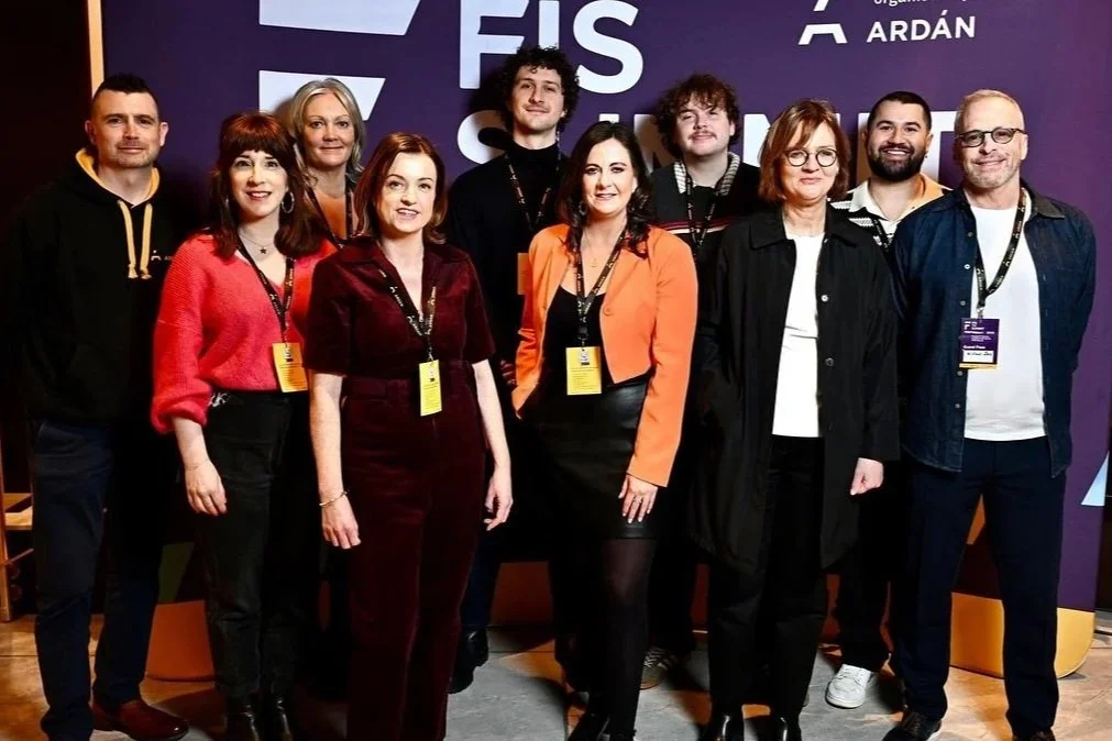Group of ten people standing in front of a purple backdrop with event information, posing for a photo at a professional conference or event, wearing conference badges.