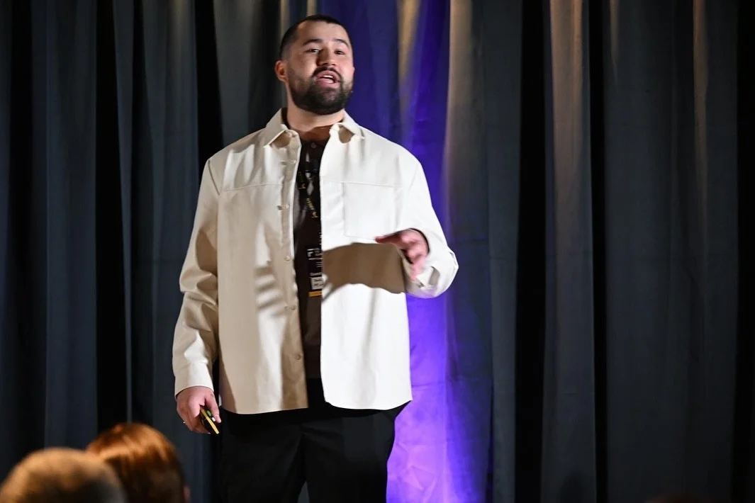 A man with a beard and dark hair speaking on stage in front of a curtain with purple lighting.
