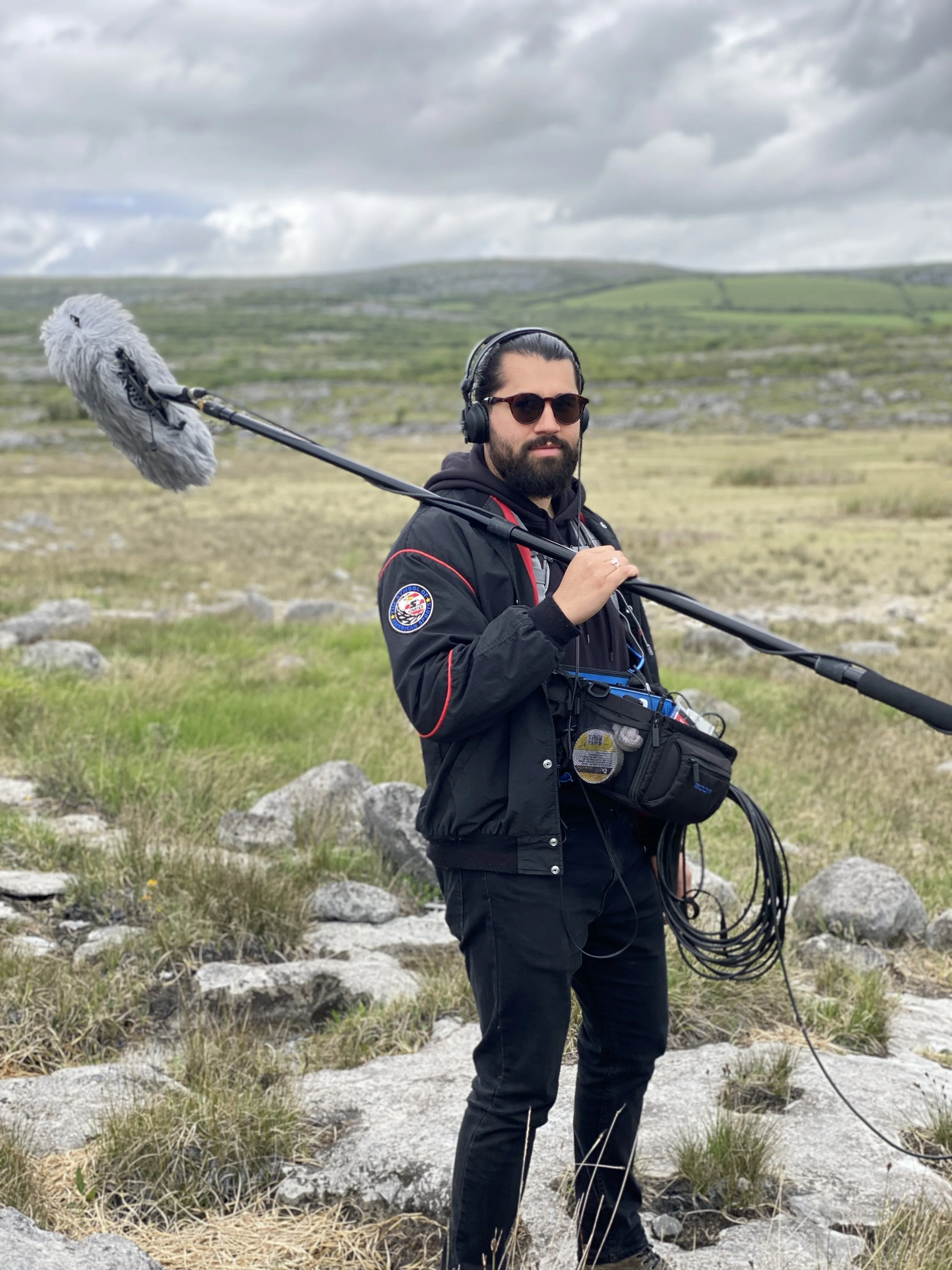 A man with dark hair and beard, wearing sunglasses, headphones, a black jacket, and black pants, holds a boom microphone outdoors on a rocky and grassy landscape with rolling hills and cloudy sky in the background.