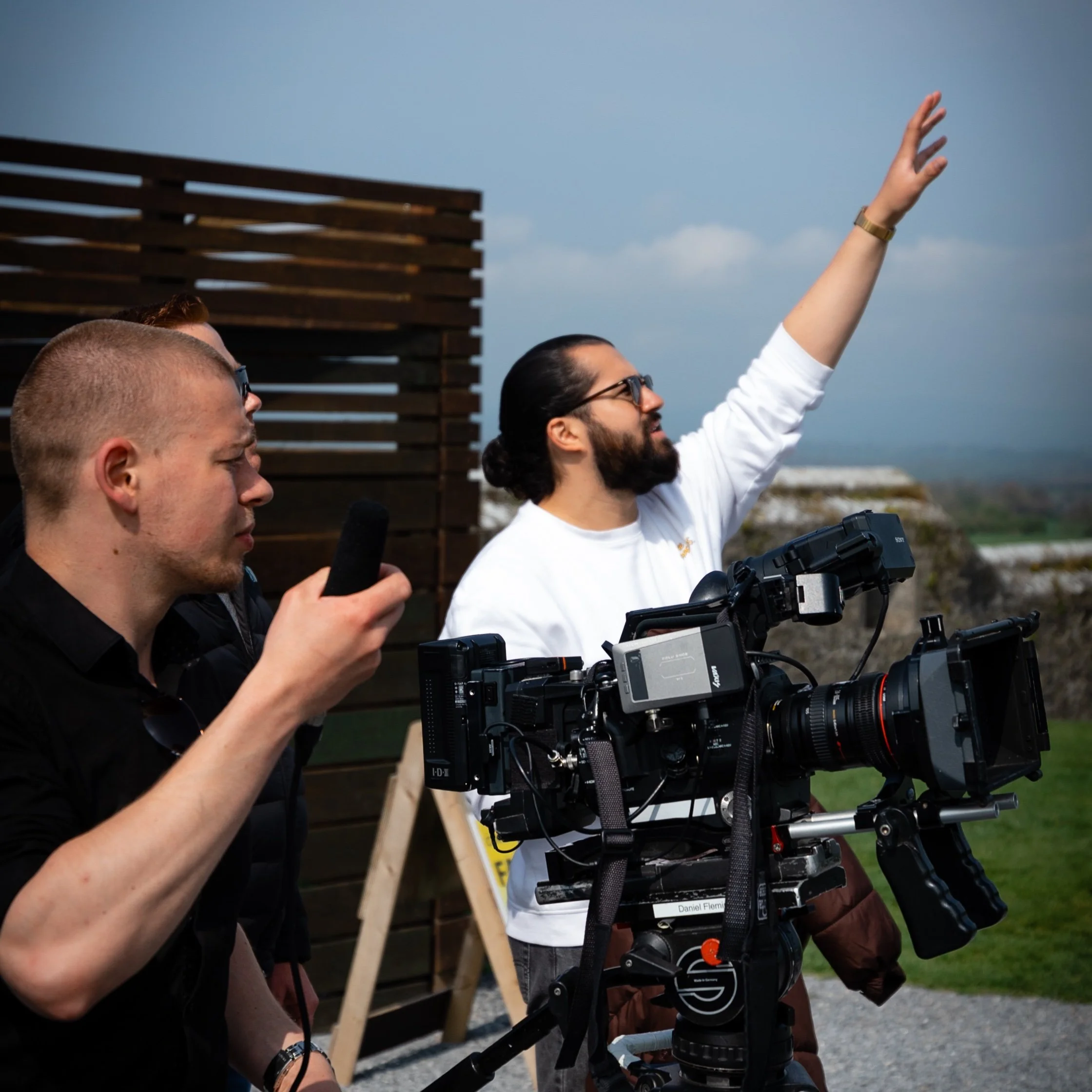 Two men operating a professional camera mounted on a tripod outdoors, with one man reaching out with his right arm and wearing glasses and a white shirt, and another man holding equipment, all against a backdrop of a blue sky and wooden structure.
