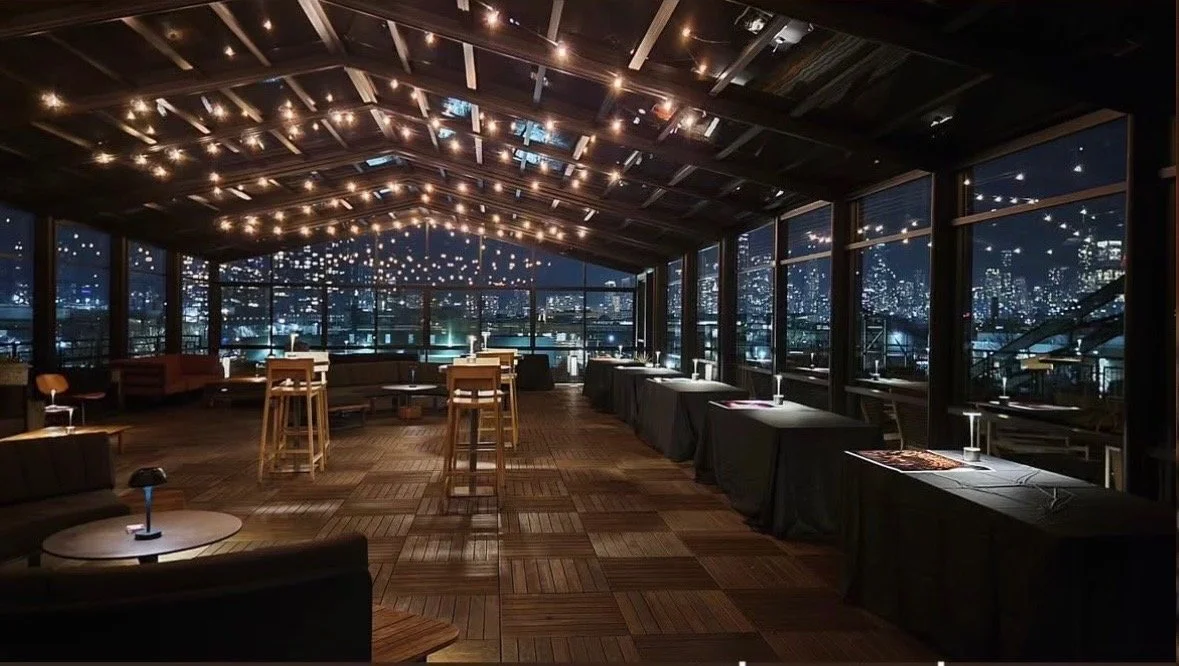 Interior of an upscale rooftop lounge at night with string lights on the ceiling, high tables, and a city skyline view through large windows.