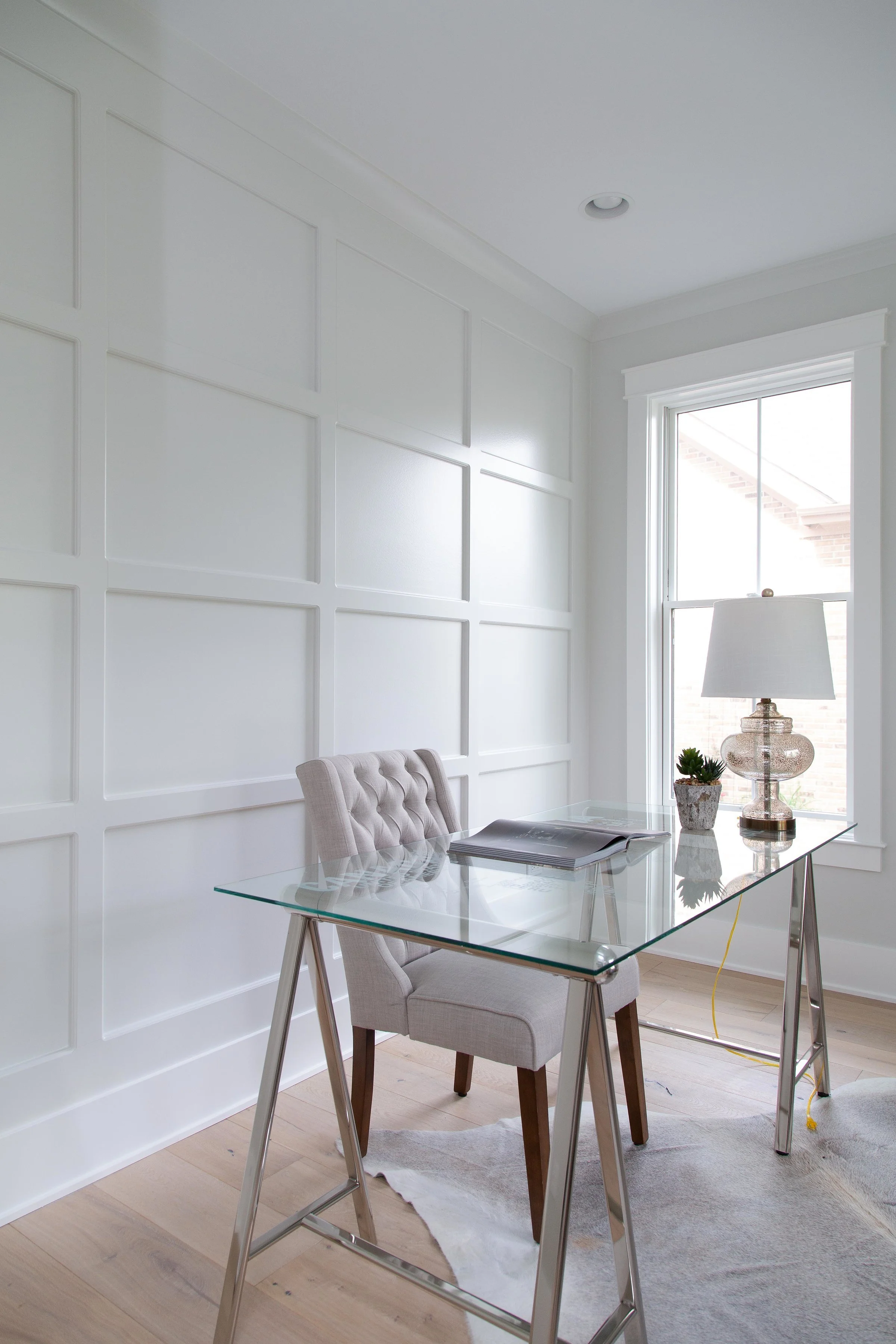 A bright, modern dining or work area with a glass table, beige upholstered chair, and decorative lamp with plants on a windowsill. The room has white paneled walls, a large window, and light wood flooring, with some wires visible near the window.