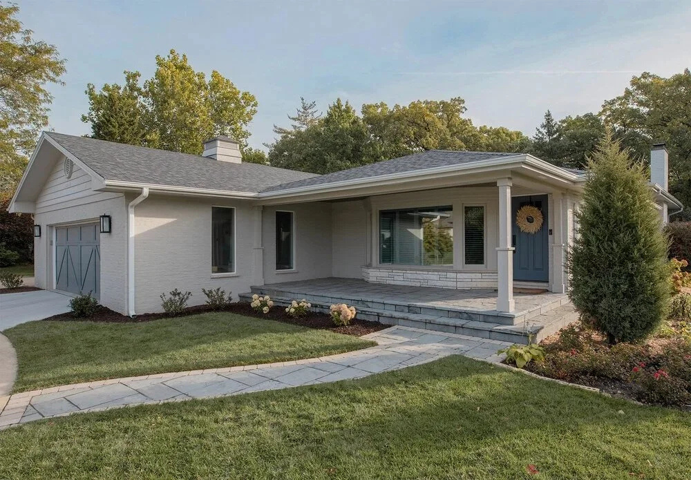Front view of a single-story house with light gray siding, a covered porch, and a blue front door, surrounded by a well-maintained lawn, bushes, and trees.