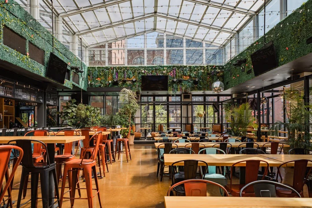 Interior of a brightly lit restaurant with a high glass ceiling, green wall decorations, and multiple seating arrangements including high tables with bar stools and standard tables with chairs, decorated with plants and modern decor.