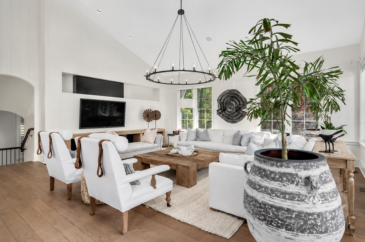 A spacious, bright living room with white walls and ceiling, polished wood floors, and large windows. The room features a large white sectional sofa, two white armchairs with brown leather straps, a rustic wooden coffee table, and a large patterned c