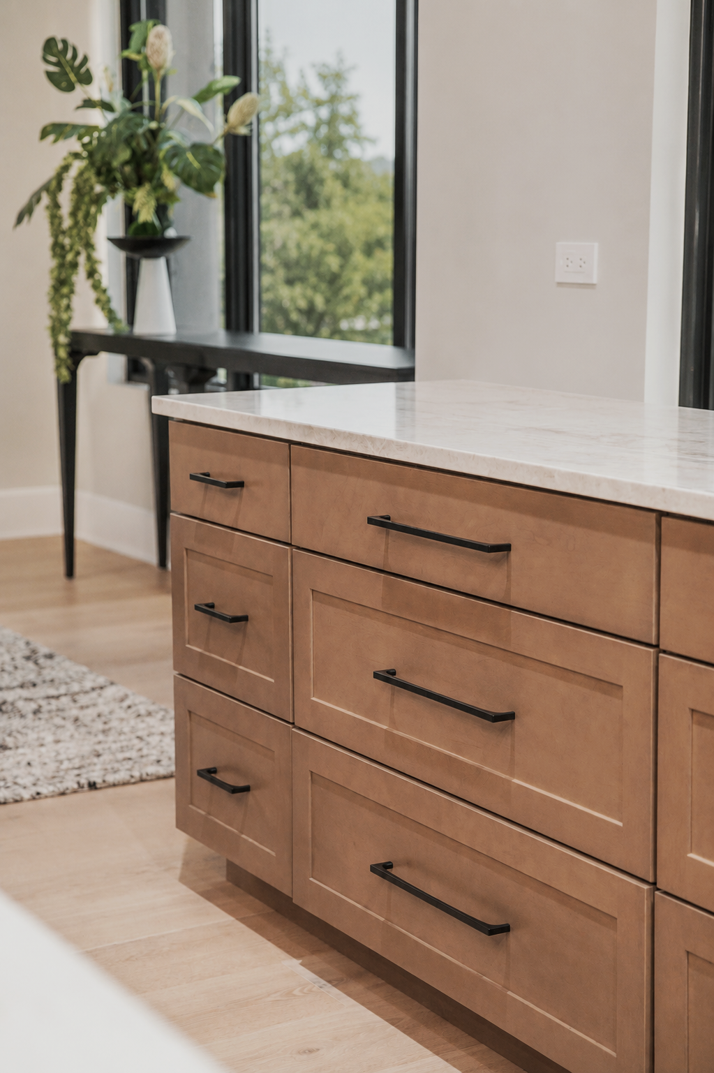 A modern kitchen with a wooden cabinet topped with a white marble countertop, a black metal handle, and a black table with a floral arrangement and a window showing green trees outside.