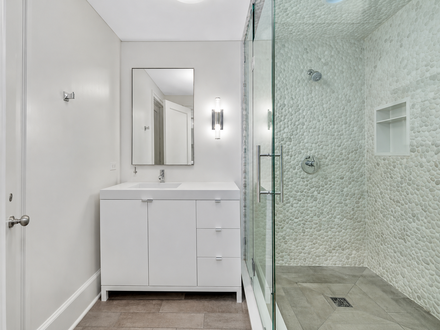 Modern bathroom with white vanity and a mirror, adjacent to a glass shower enclosure with pebble wall design and built-in shelf.