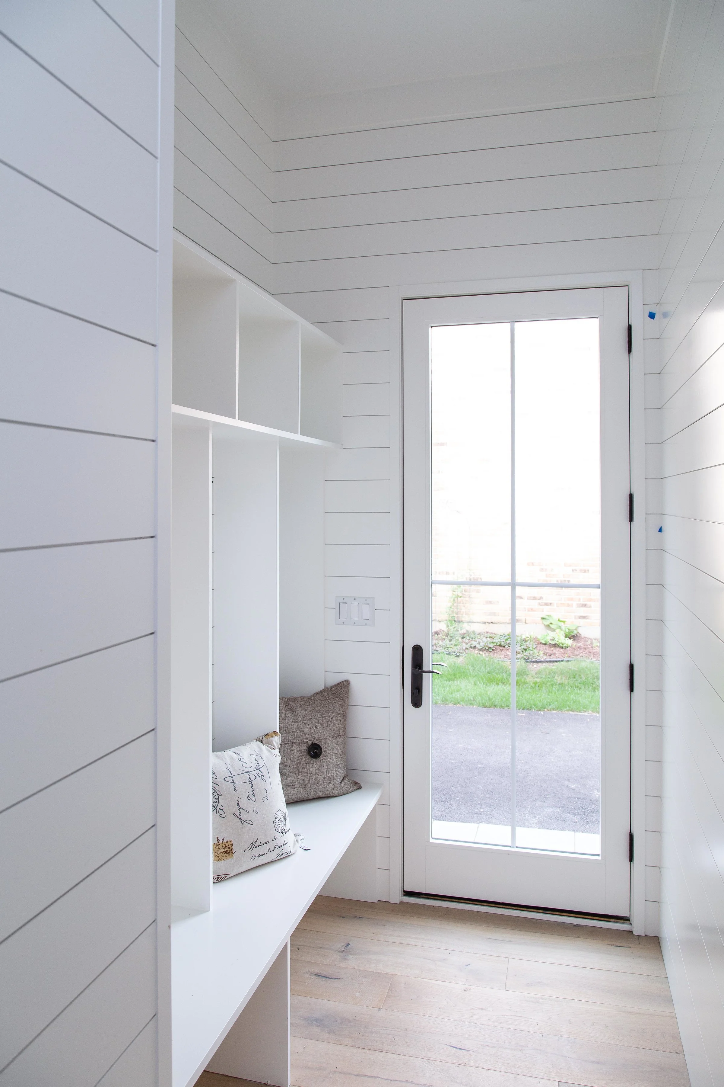 A bright room with white shiplap walls and ceiling, a sliding glass door leading outside to a grassy yard, with a small bench and decorative pillows inside.