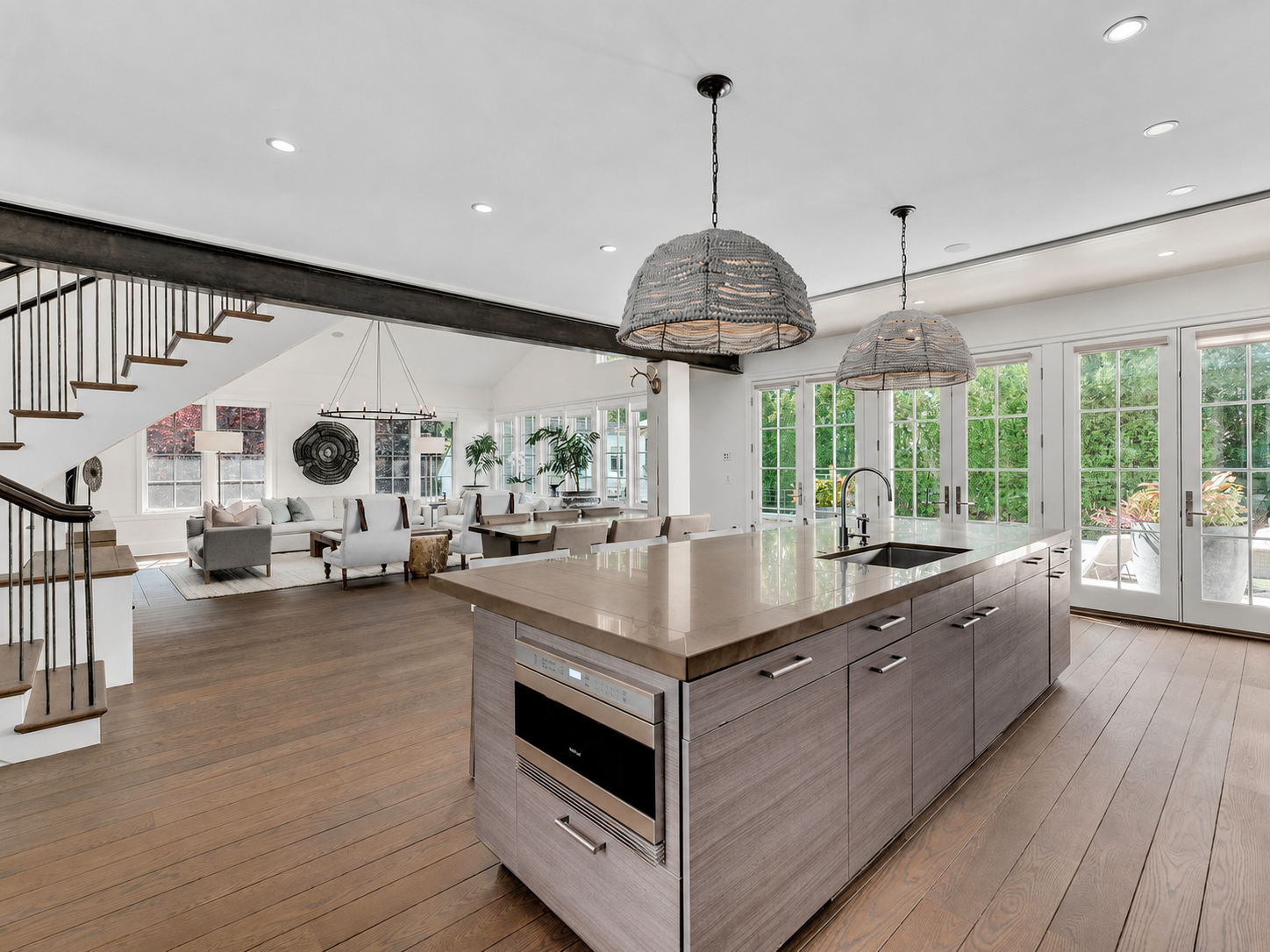 Open-concept living room and kitchen with large windows, pendant lights, wooden flooring, and white walls.