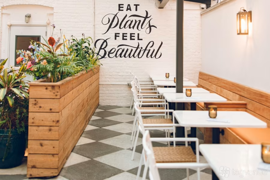 Empty outdoor cafe with white tables, wooden benches, and chairs, decorated with small candles, next to a large wooden planter filled with colorful flowers and greenery. A white brick wall features a quote: 'Eat plants feel beautiful'.