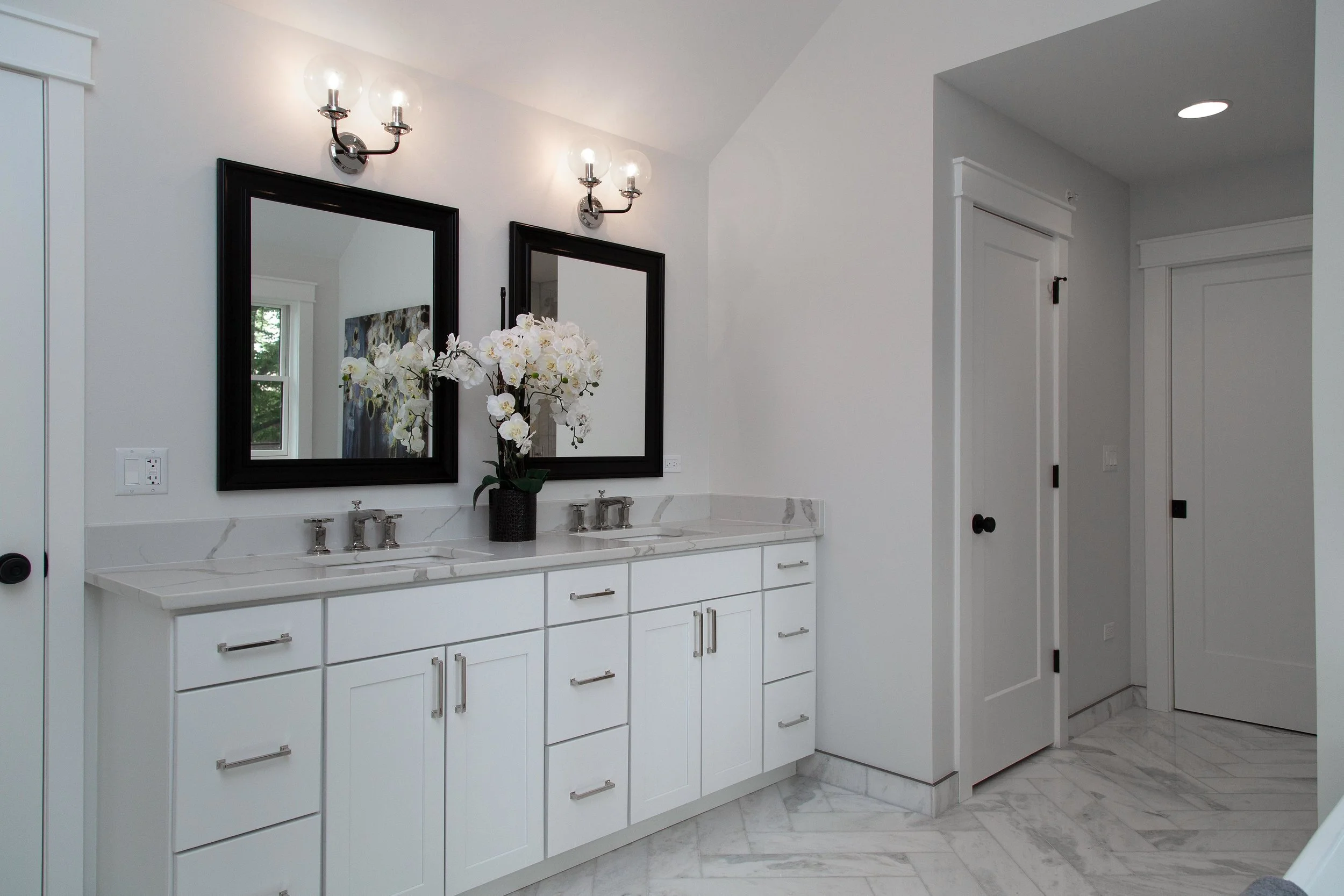 A modern bathroom with white cabinets, marble countertops, two black-framed mirrors, a white orchid plant, and wall-mounted light fixtures with bulb-style bulbs. There is a window reflected in one of the mirrors and a door on the right side.
