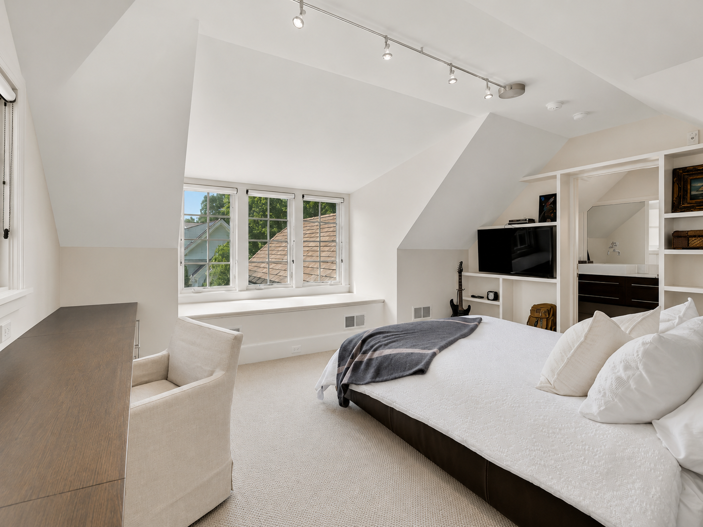 A bedroom with a white bed, beige carpet, a wooden desk, cream armchair, large window with a view of neighboring houses, a TV, guitar, and a small sink area visible through a doorway.