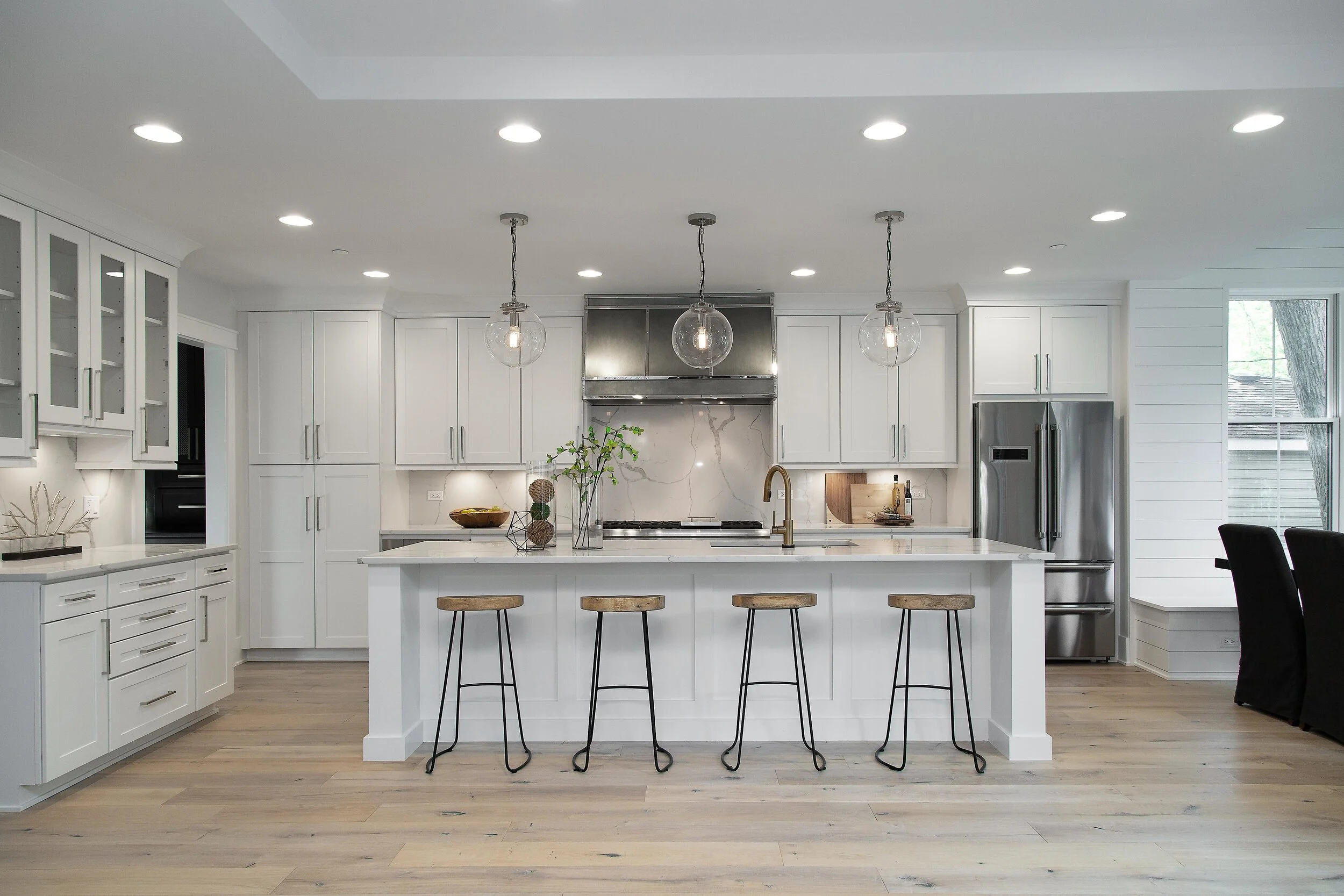 Modern white kitchen with island, wooden stools, pendant lights, stainless steel appliances, and large window.