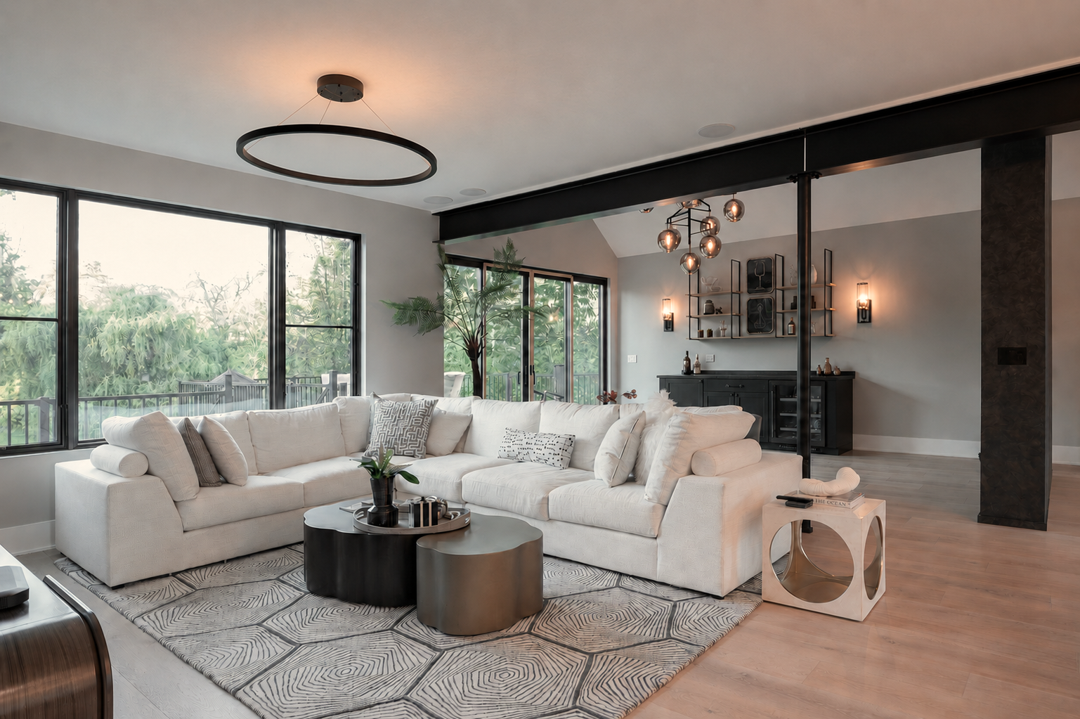 Modern living room with large white sectional sofa, black and gray coffee tables, large window with greenery outside, indoor potted plant, and contemporary decor lighting.