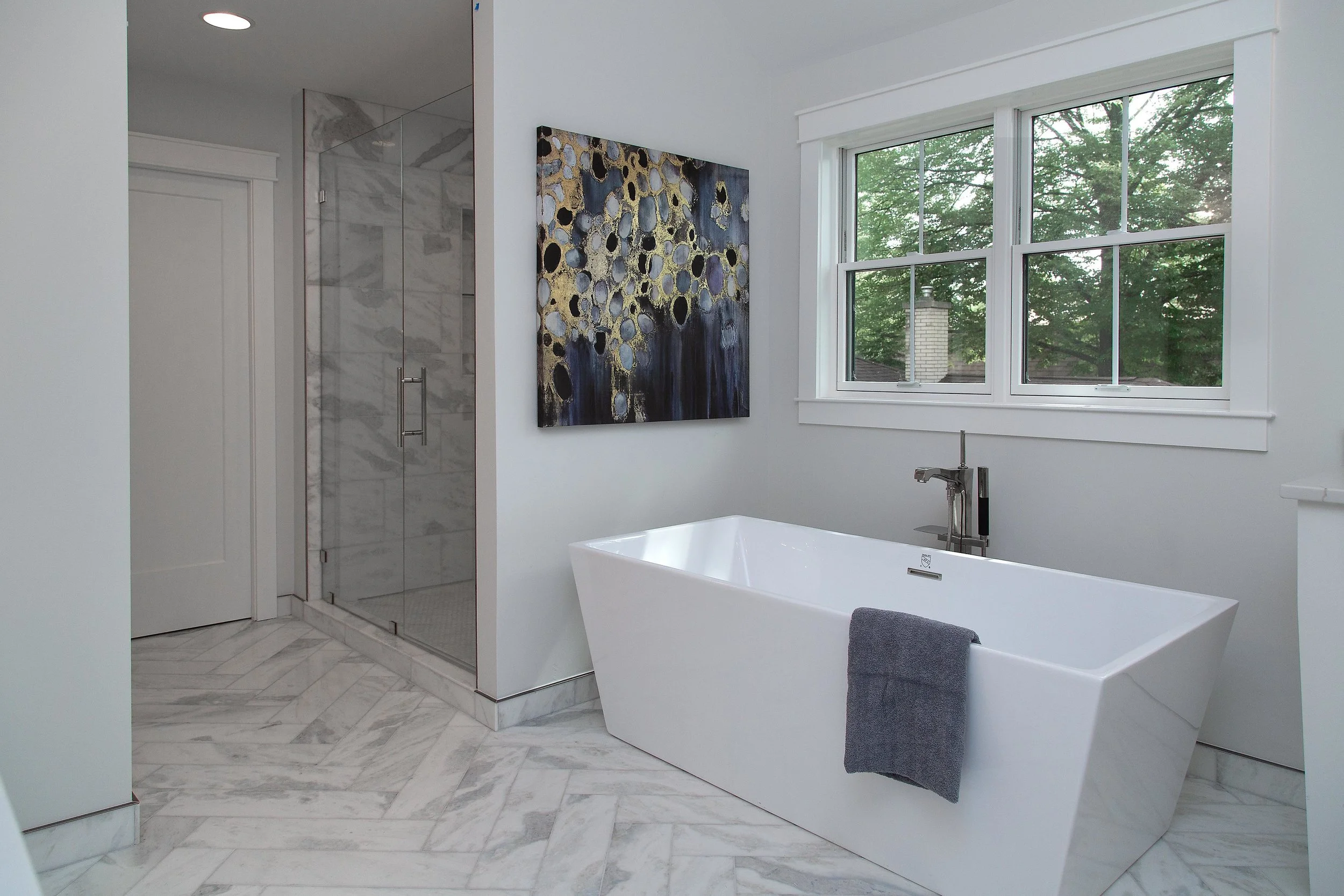 Modern bathroom with a bathtub, a window with green trees outside, abstract wall art, and marble flooring.