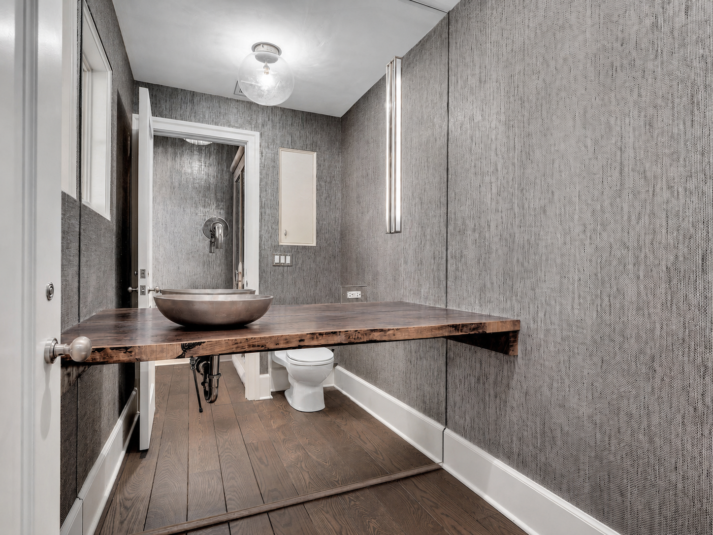 Modern bathroom with gray textured walls, wooden floor, a floating wooden vanity with a metallic vessel sink, and a partially visible toilet beneath the vanity.