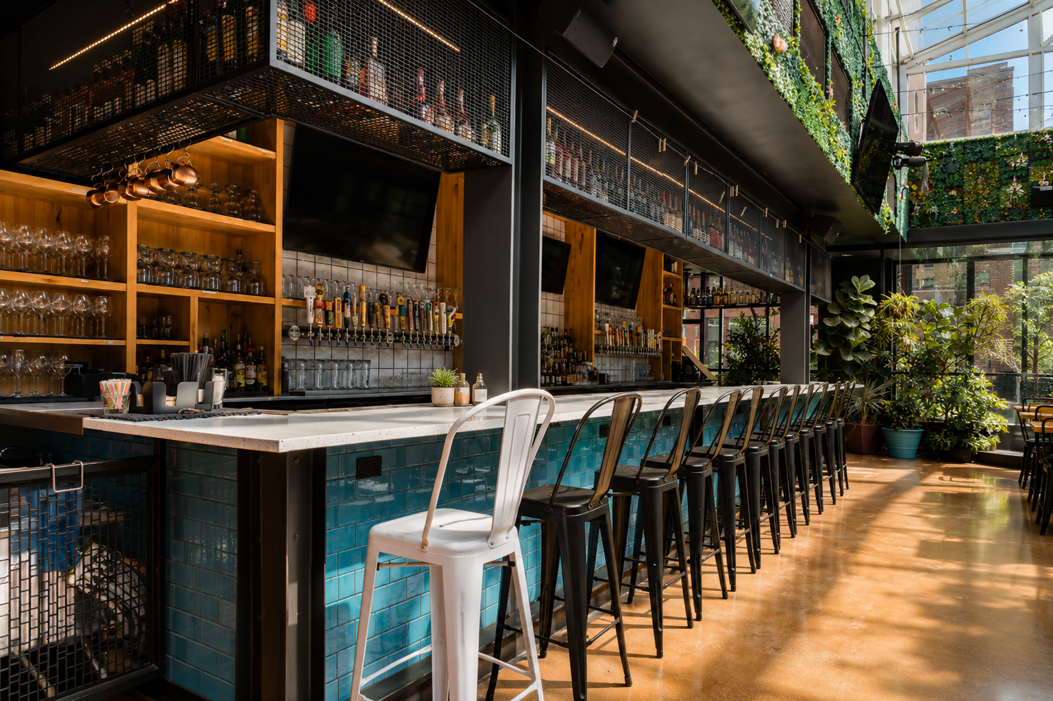 A modern bar with a white countertop, black and white chairs, potted plants, and large windows allowing natural light to fill the space. The bar has wooden shelves with bottles and glasses, and a decorative green wall with plants and hanging lights.
