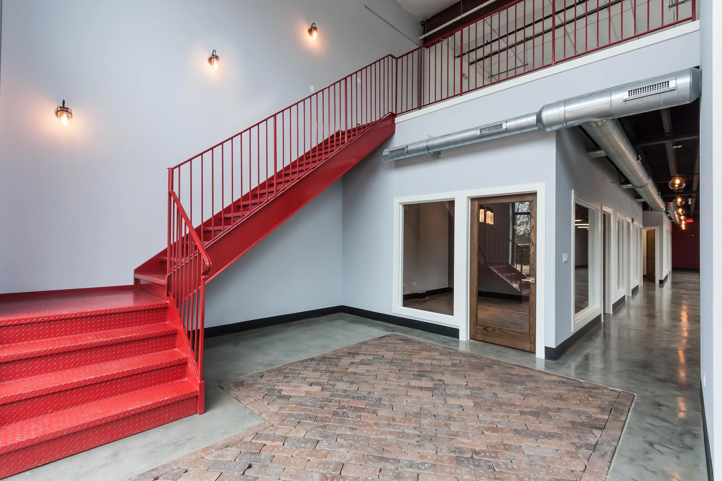 Interior of a modern building with a red staircase, a brick and concrete floor, gray walls, and exposed silver ductwork on the ceiling. There are wall-mounted light fixtures illuminating the space.