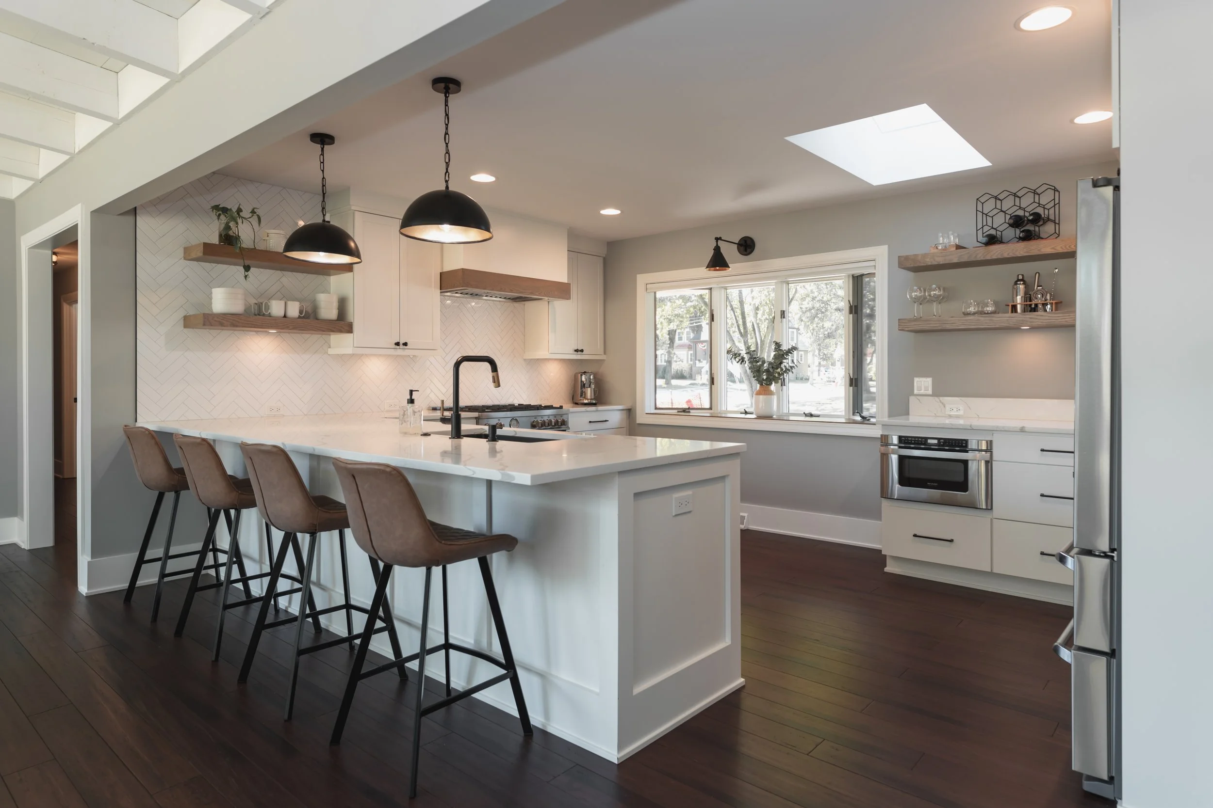 Modern kitchen with white cabinets, marble island, black pendant lights, and large window overlooking trees.