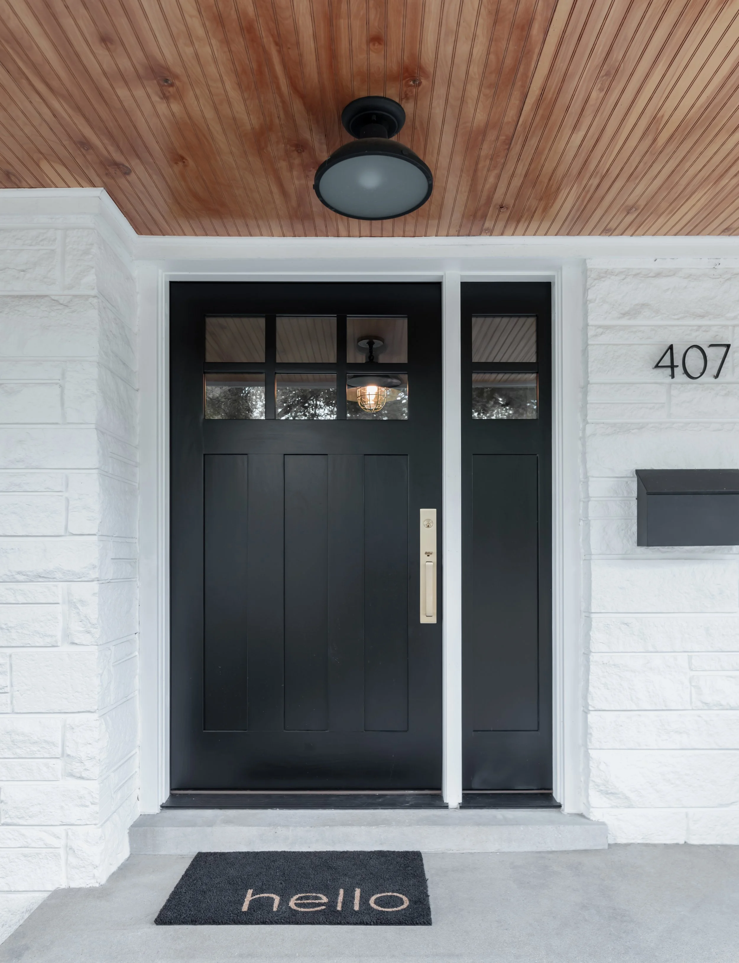 Front porch with a black door, white brick walls, a black exterior light, and a doormat with the word "hello" on a cement porch.