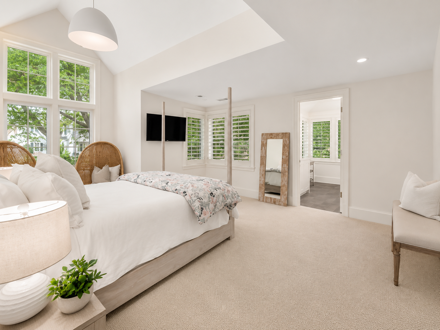 Bright bedroom with large windows, white walls, beige carpet, and a bed with white linens, rattan headboard, and floral blanket. There is a bedside table with a lamp and plant, and a mirror leaning against the wall near an adjoining bathroom with shi