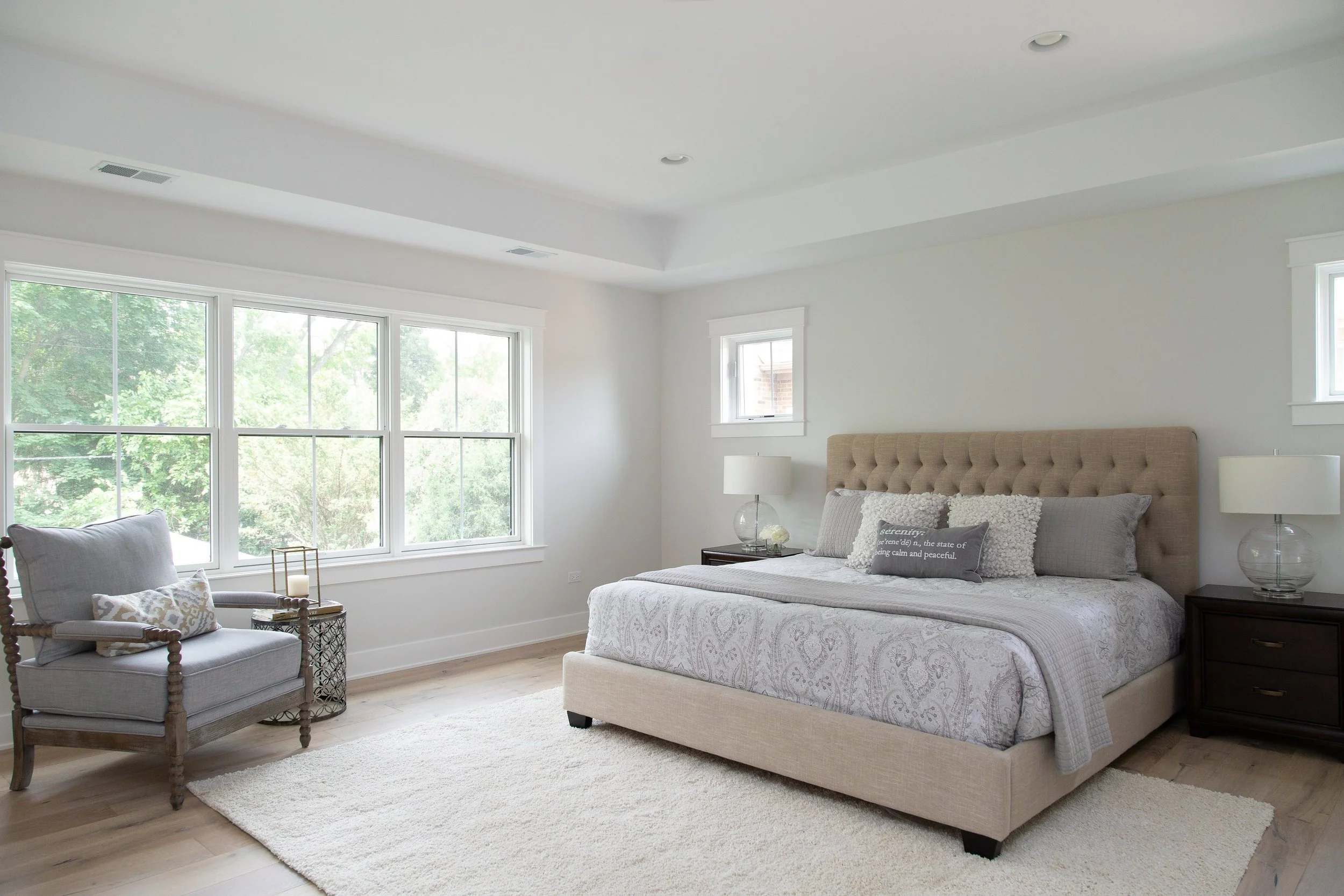 A bright and airy bedroom with large windows showing green trees outside. It features a beige upholstered bed with patterned bedding, matching pillows, two dark wooden nightstands with glass lamps, a white armchair with decorative pillows, a beige ar