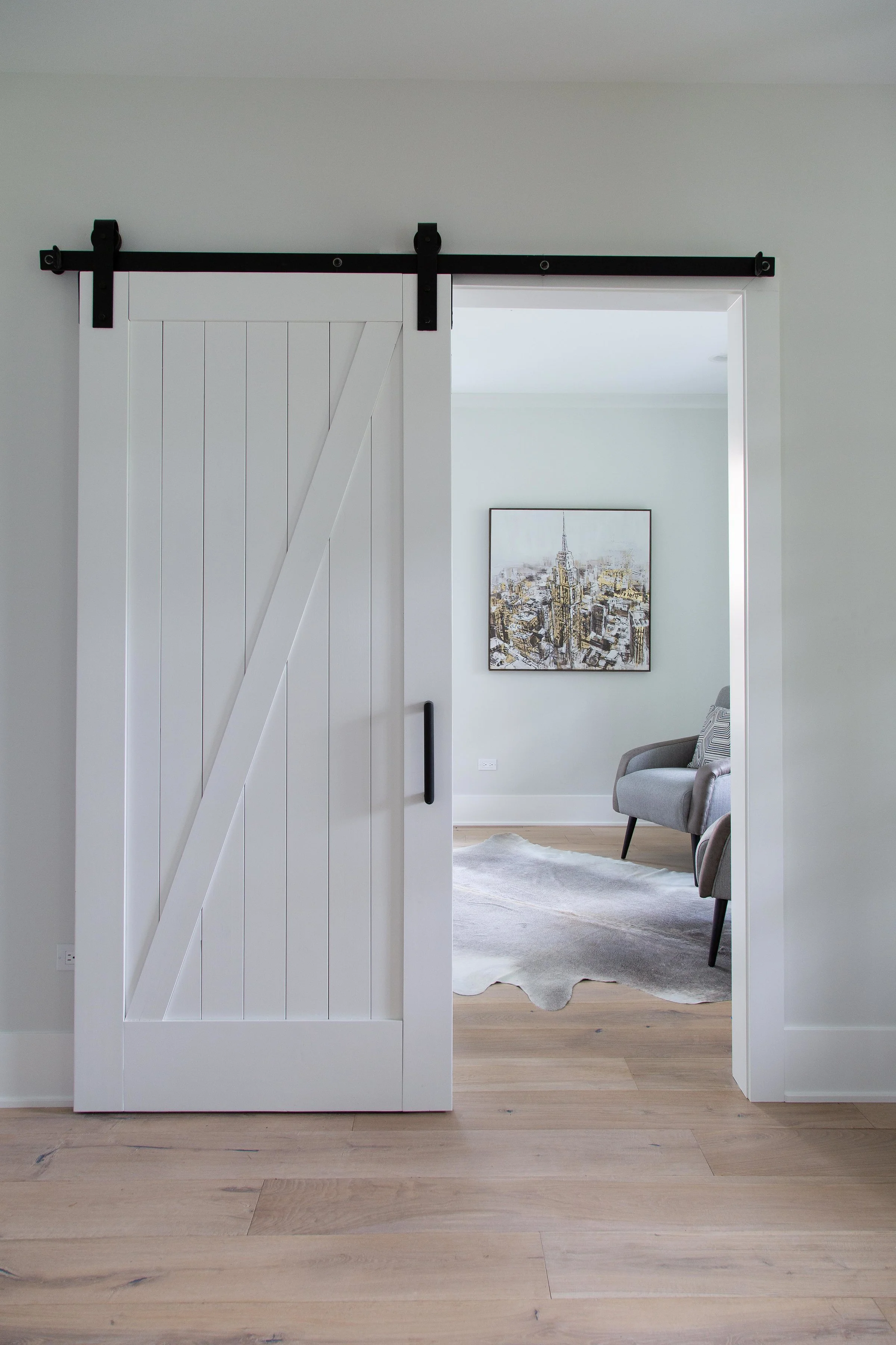 Interior view through a sliding barn door, revealing a living room with a chair, a painting of the New York City skyline, a light-colored rug, and hardwood flooring.