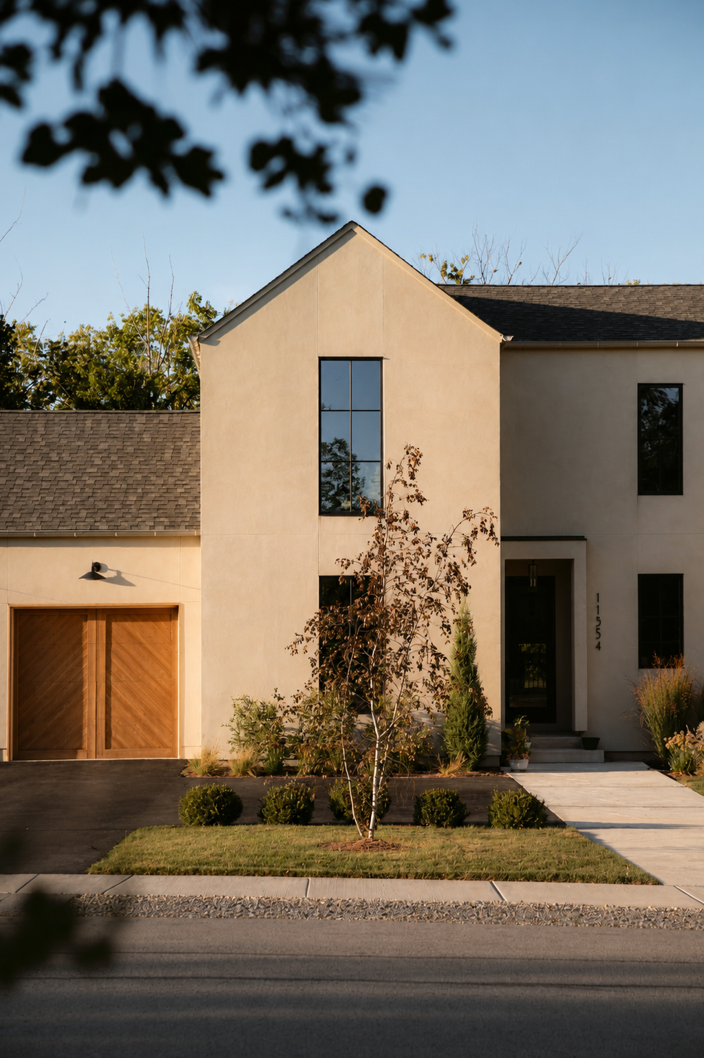 Modern two-story house with beige stucco exterior, black trim, and a wooden garage door, front yard with small tree and shrubs, sidewalk, and driveway, under clear blue sky.