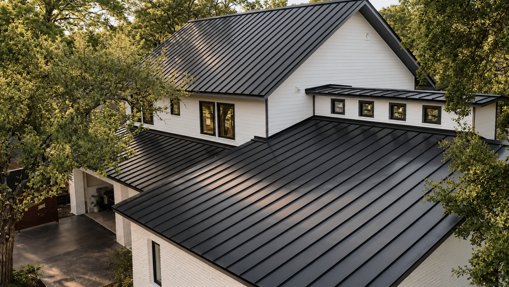 A modern house with white siding and black metal roofing, surrounded by green trees.