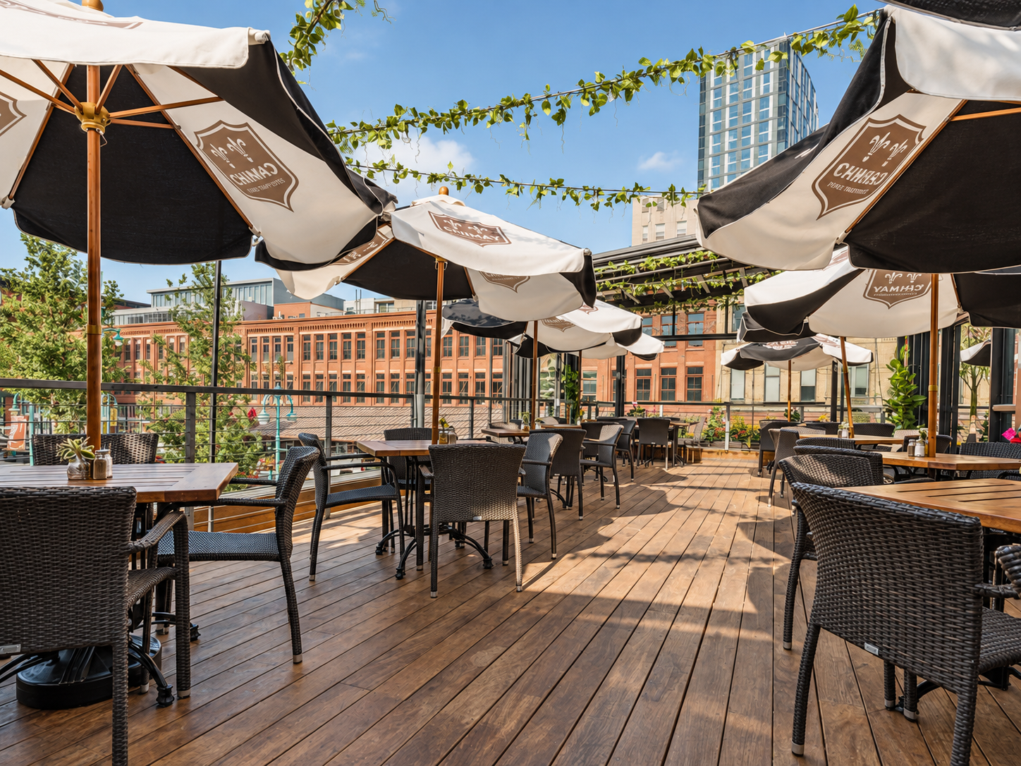 Outdoor rooftop restaurant with tables, chairs, and large umbrellas on a sunny day, with a cityscape in the background.