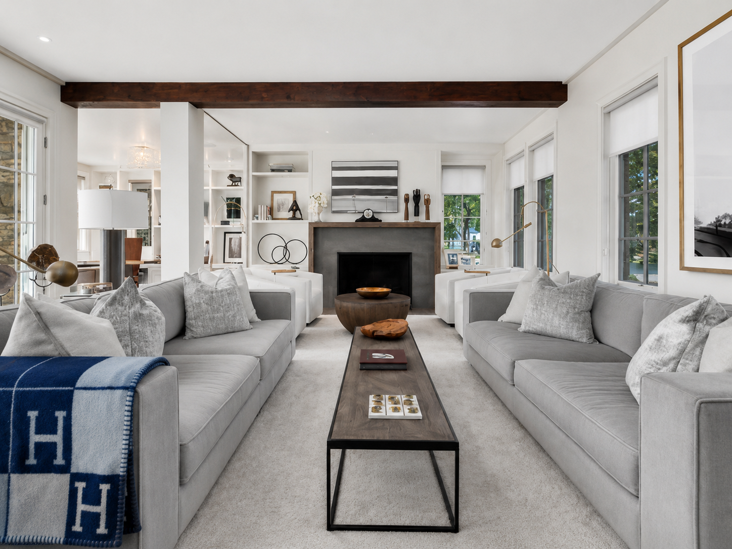 A modern, bright living room with two gray sofas, decorative pillows, a dark wooden coffee table, and a fireplace surrounded by shelving with artwork and decor. Large windows let in natural light.