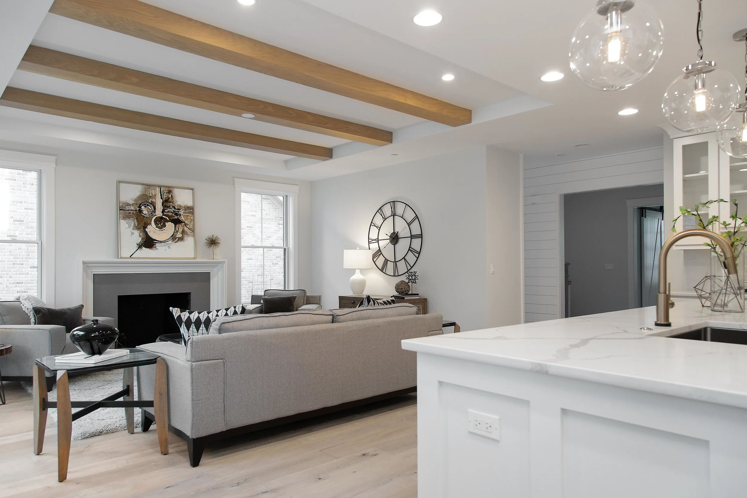 Modern living room with gray sofa, abstract artwork above fireplace, large wall clock, and two windows. Kitchen island with brass faucet and pendant lights in the foreground.
