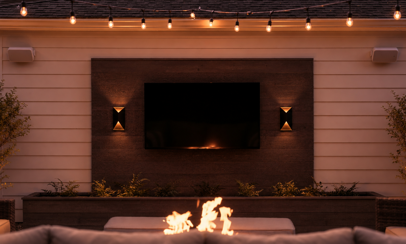 An outdoor patio setup at sunset with string lights overhead, a mounted flat-screen TV on a dark wooden accent wall, two wall-mounted lights on either side of the TV, a fire pit, and lush plants in a planter box.