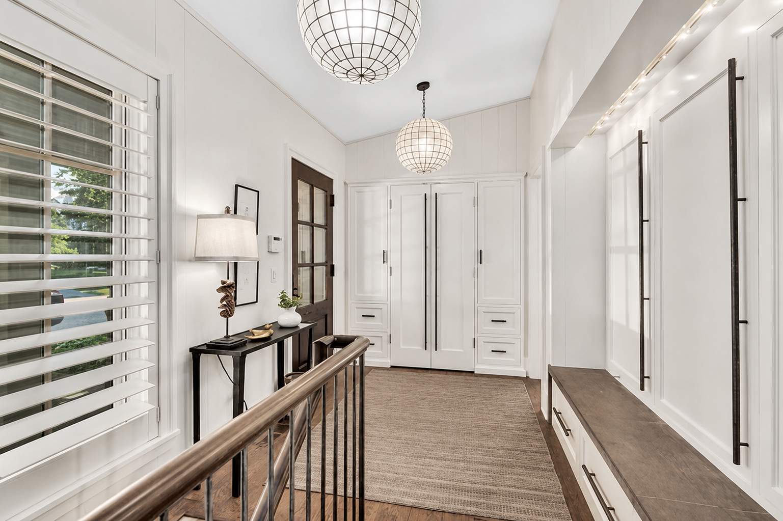 Bright hallway with white walls, wooden flooring, an area rug, built-in storage with black hardware, a wooden bench, a window with white blinds, and modern pendant lights.