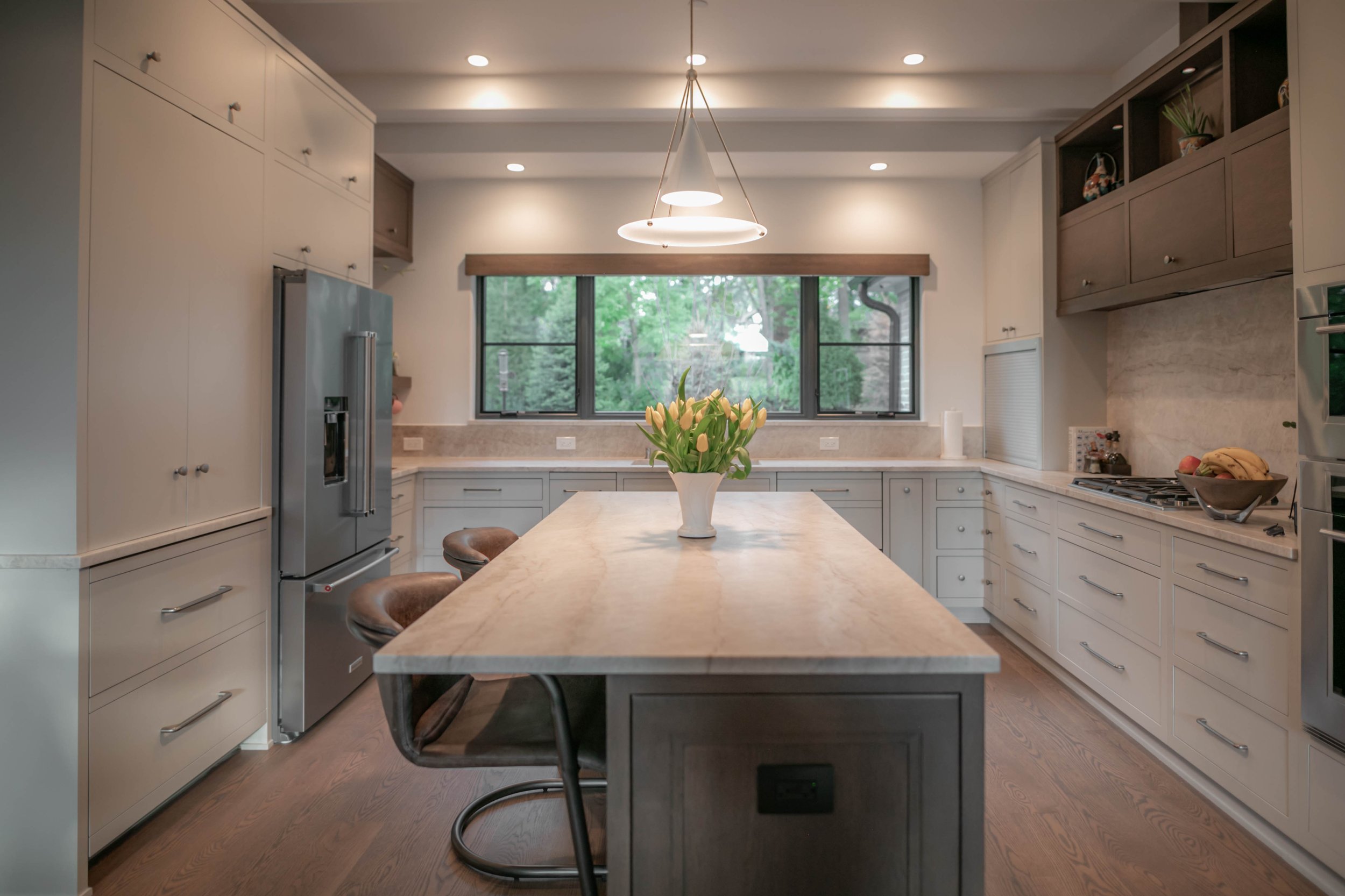 Modern kitchen with white cabinets, a central island with a flower vase, stainless steel refrigerator, and large window with greenery outside. There are three brown bar stools around the island and a hanging light fixture above.