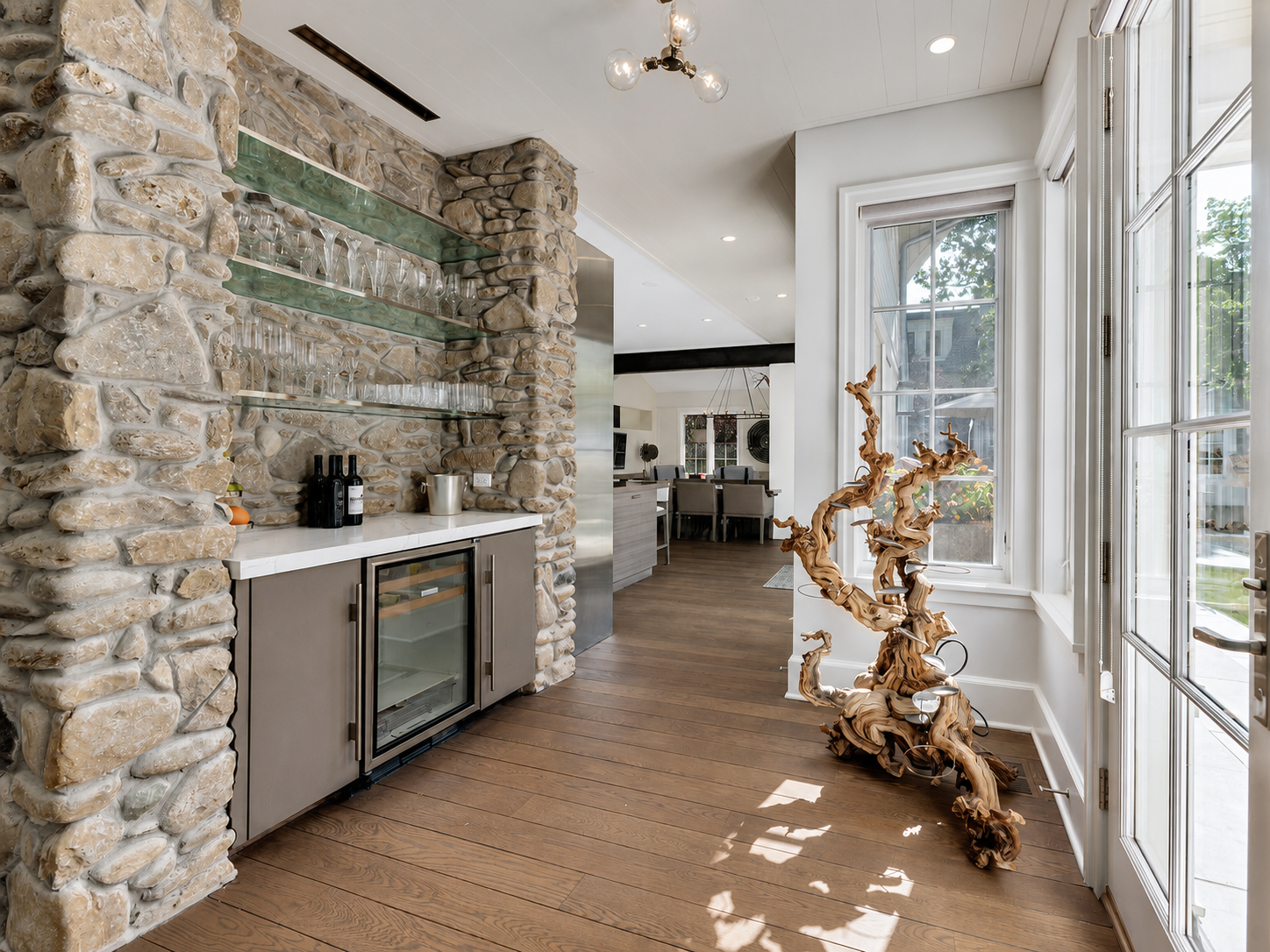 Interior of a modern kitchen with a stone accent wall, glass shelves with glassware, a small wine fridge, and a unique twisted wooden sculpture near large windows letting in natural light.