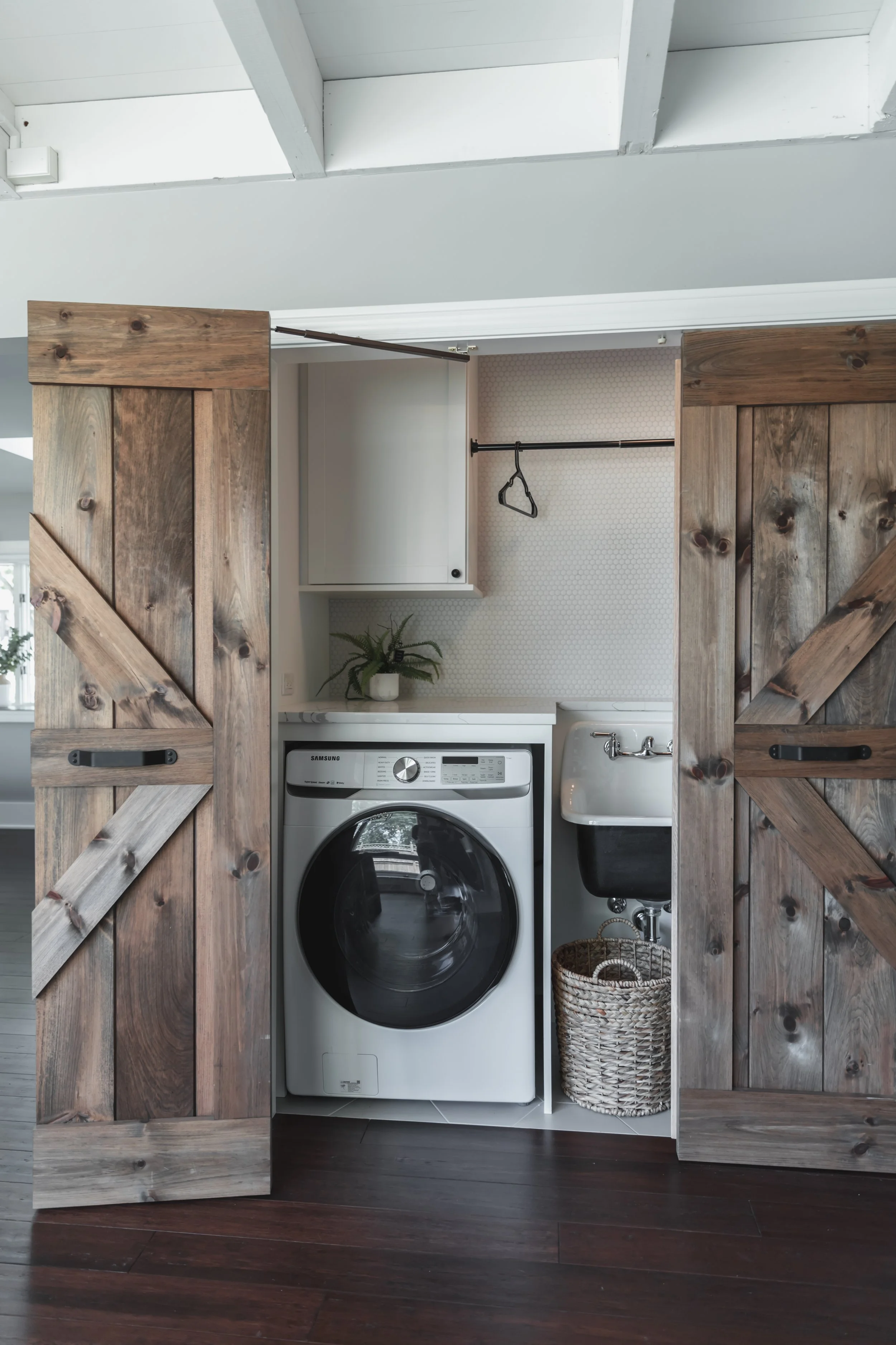 A laundry nook behind sliding barn doors made of reclaimed wood, with a front-loading washing machine, a small white cabinet, a woven laundry basket, a laundry sink, and a plant on the countertop.
