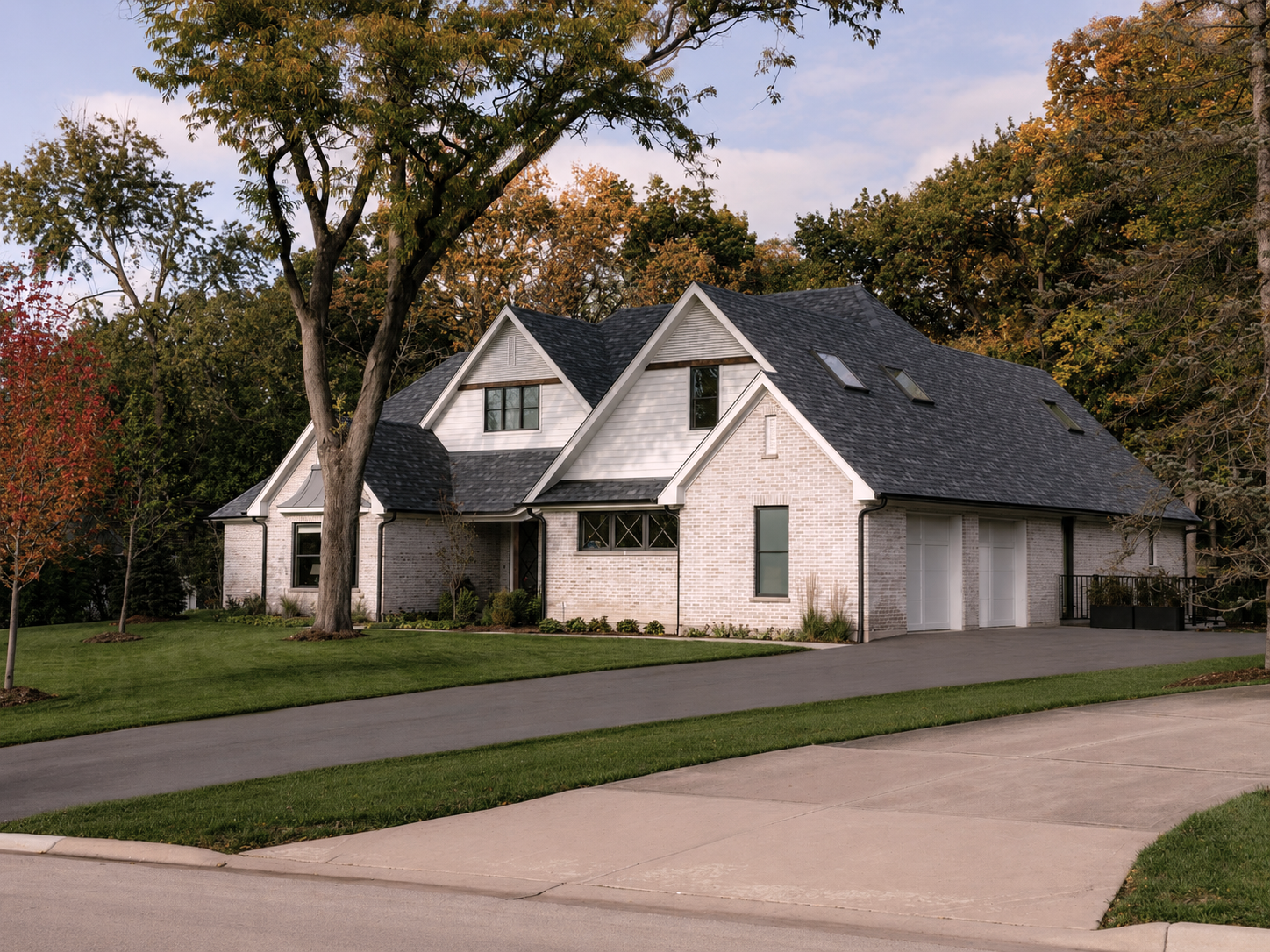 A two-story house with a light-colored brick exterior, black roof, multiple dormer windows, a driveway, and surrounded by trees with fall foliage.