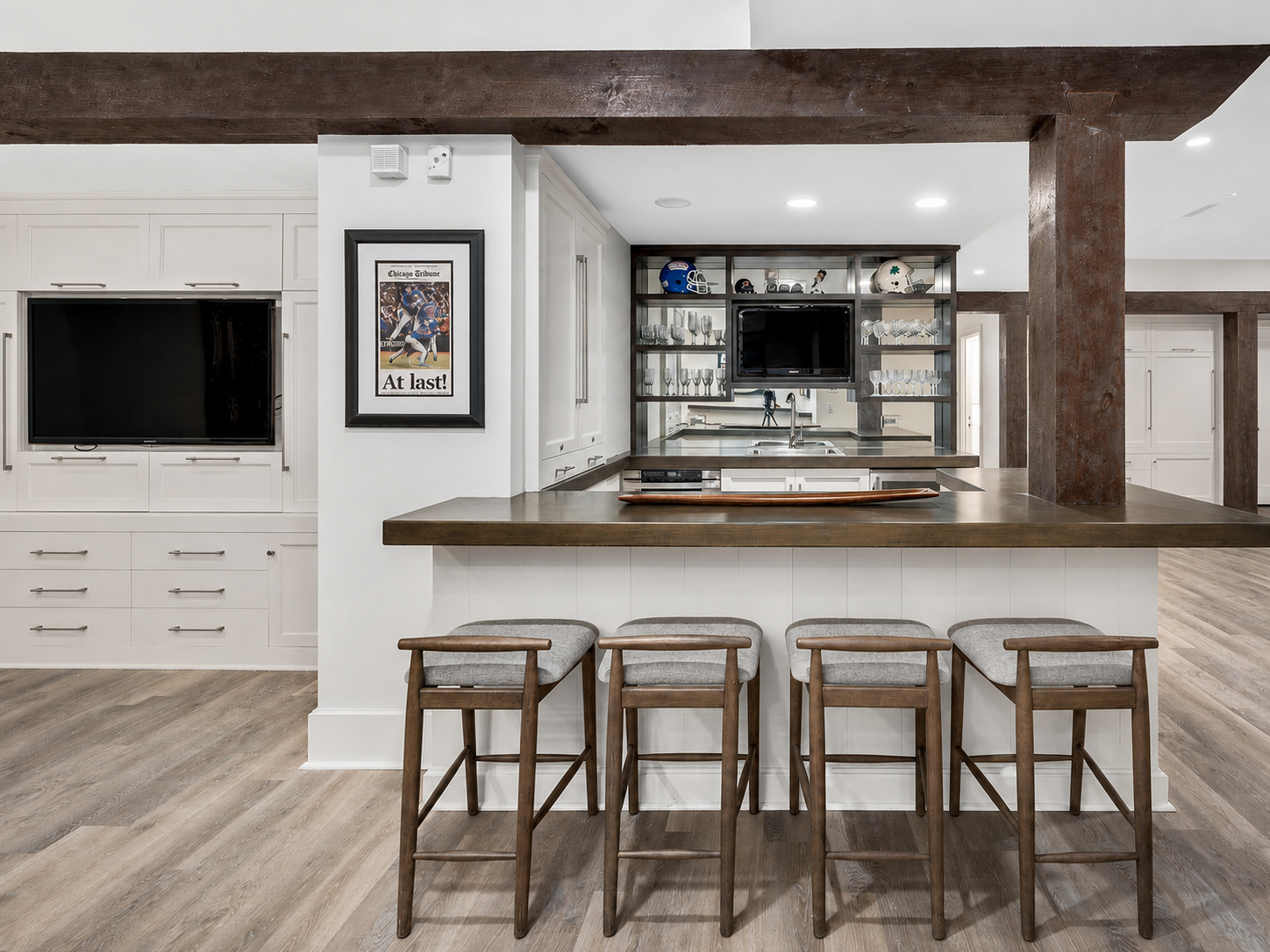 Modern kitchen with white cabinetry, a central island with a wooden countertop, four gray bar stools, and sports helmets as decor. There's a wall-mounted TV, framed sports poster, and open shelving holding glasses and small items.