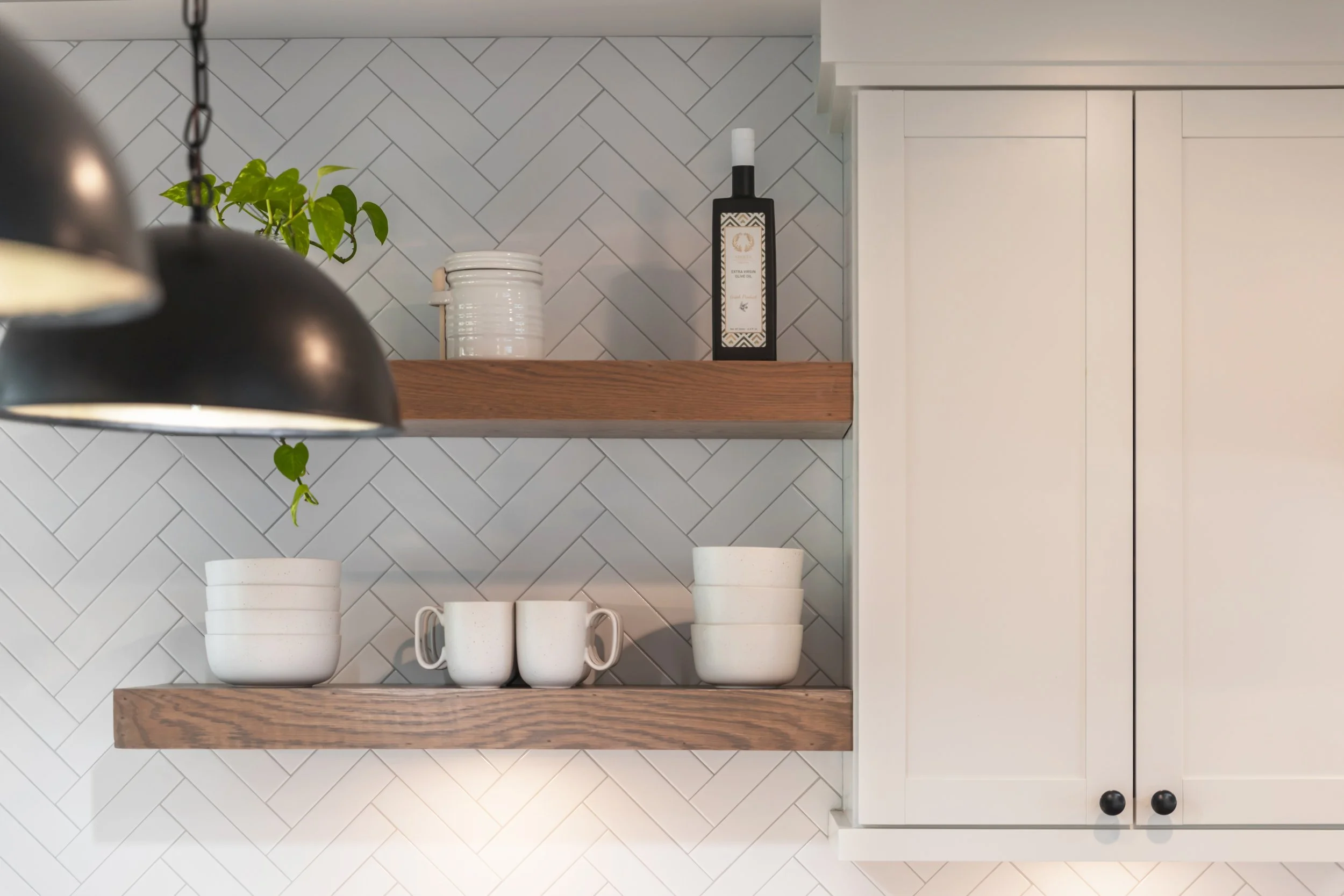 Kitchen with white cabinets, two wooden shelves, a gray chevron tile backsplash, a potted plant, white mugs, a jar, and a bottle of extra virgin olive oil.