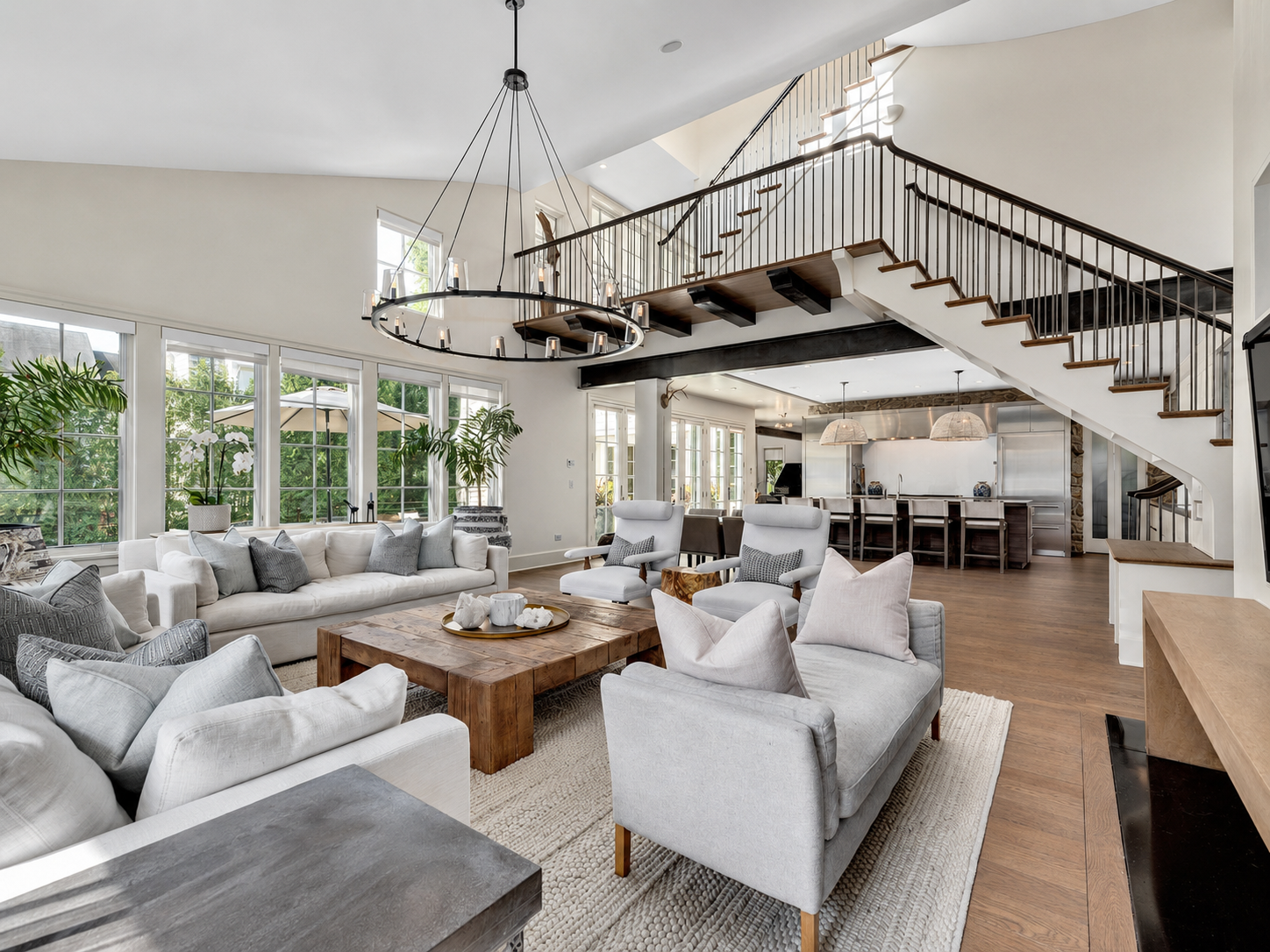 Bright, spacious living room with white sofas and armchairs, a rustic wooden coffee table, and large windows with greenery outside. An open kitchen and staircase with black railing lead to an upper floor. Modern chandelier hangs from the ceiling.