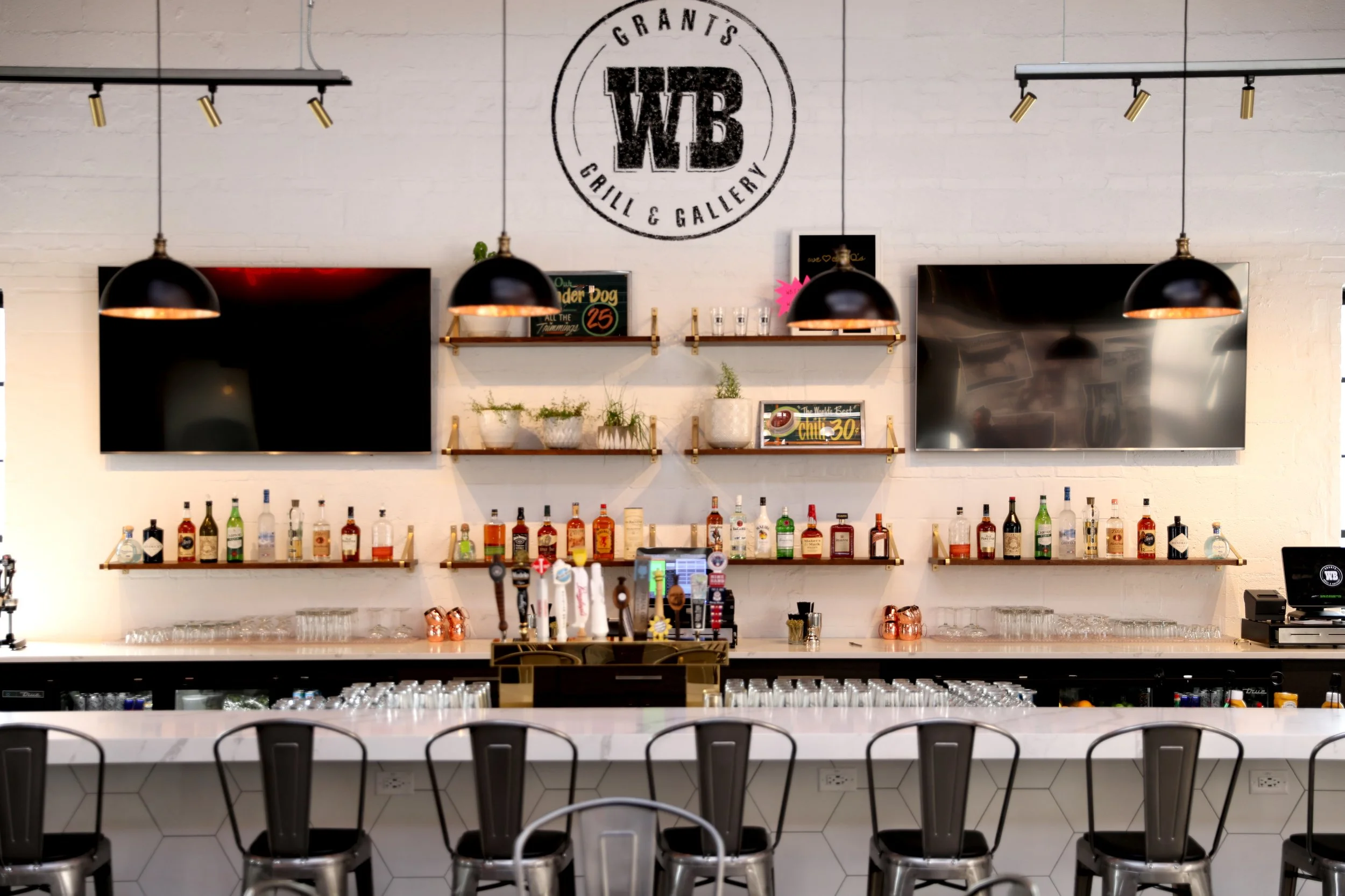 Inside view of a bar or restaurant with a white brick wall, mounted televisions, black pendant lights, shelves with plants and bottles, and bar stools at the counter.