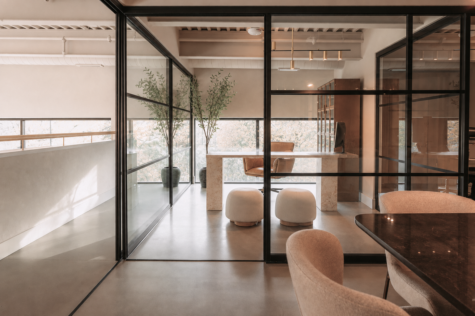 Modern office space with glass walls, a desk with a computer, leather chair, two potted plants, and seating area with ottomans.