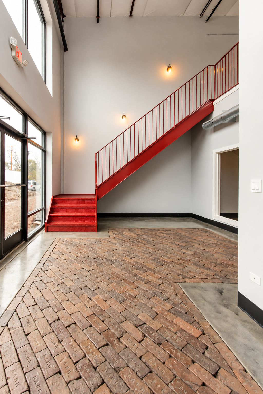 Interior of a modern building lobby with a red staircase, brick floor, large windows, and industrial-style lighting.