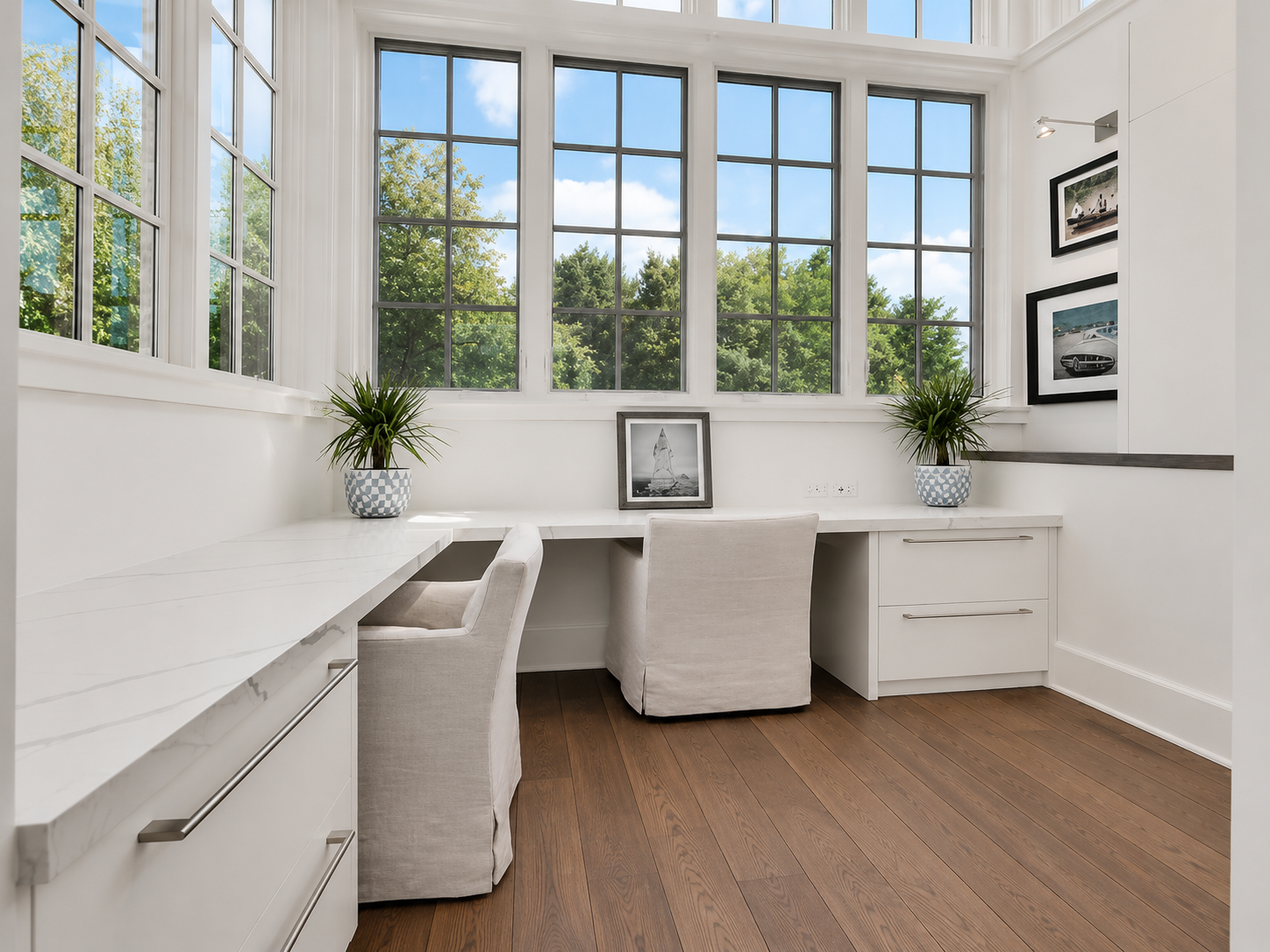 A bright corner in a modern room with large floor-to-ceiling windows showing green trees outside, white walls, a white desk with drawers, two beige upholstered chairs, framed photographs on the walls, potted plants, and a wooden floor.